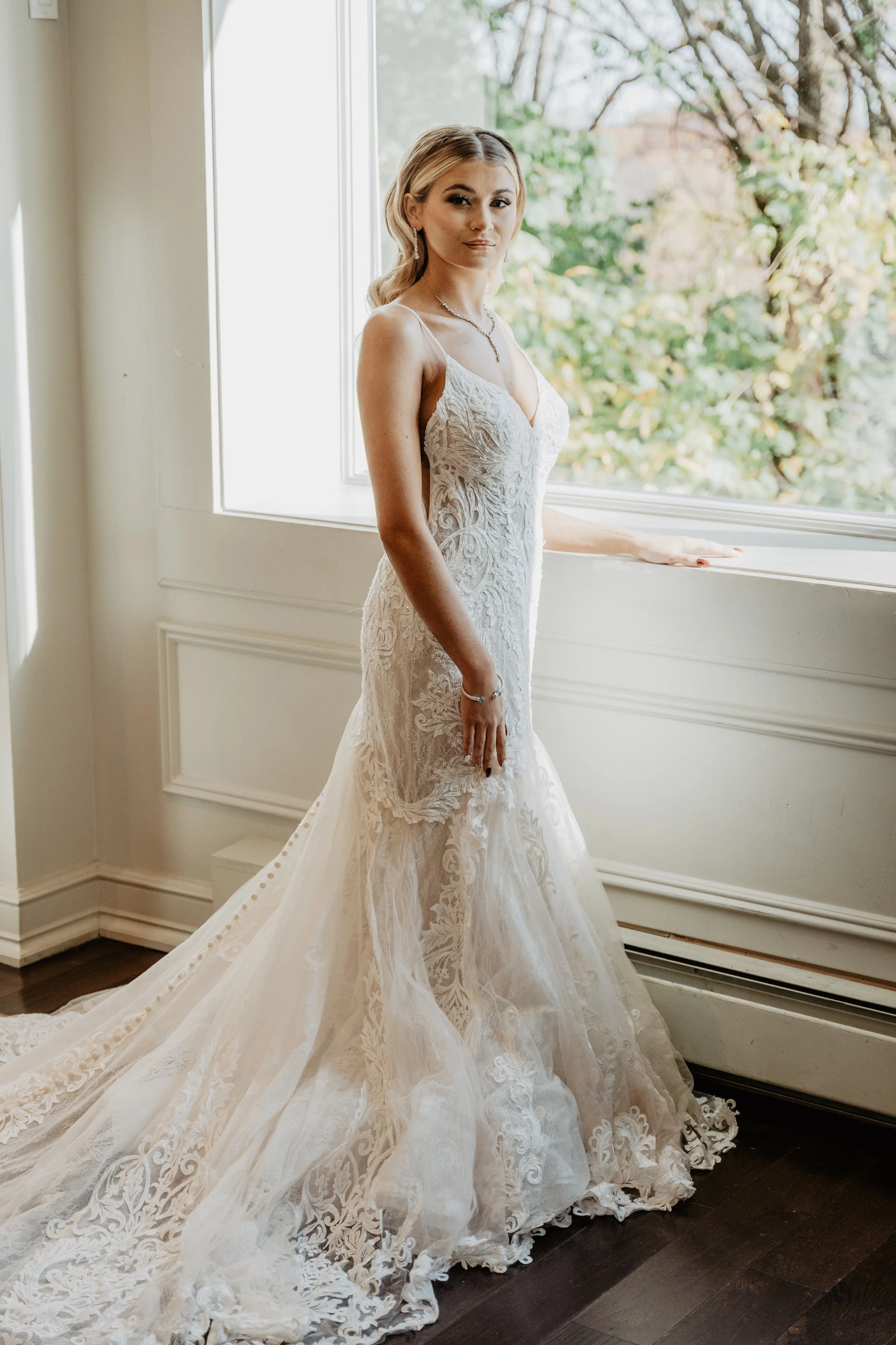 Bride in a white lace wedding gown standing by a window with autumn trees outside.