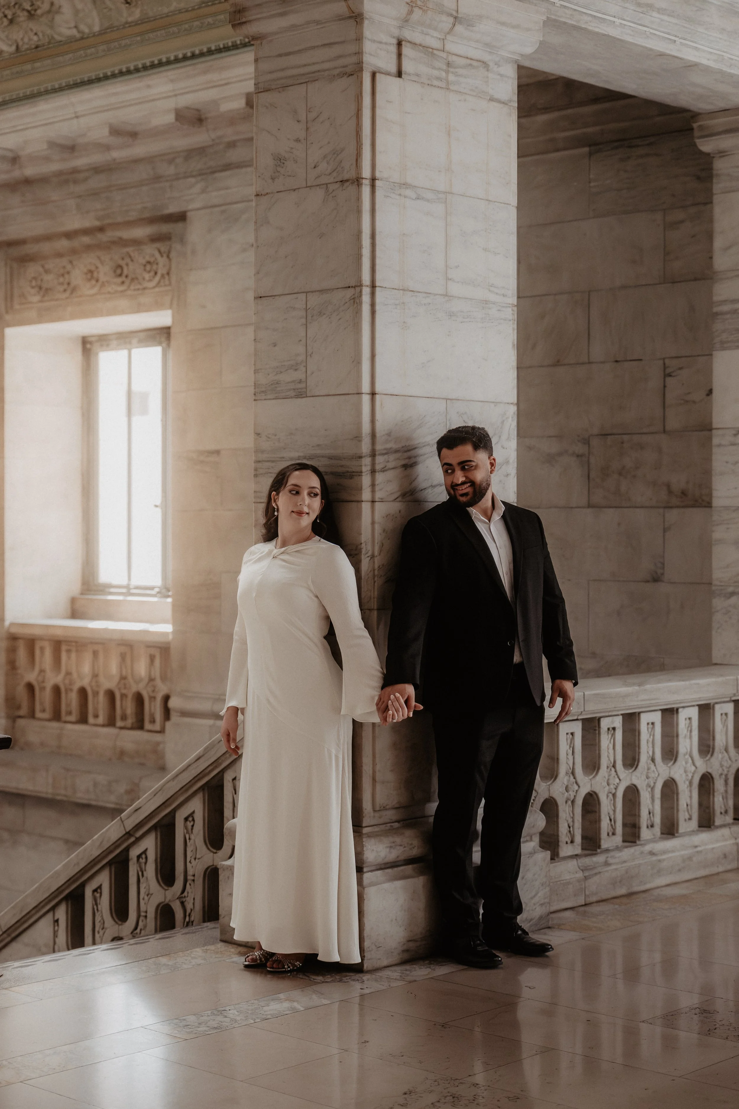 A couple dressed in formal attire, the woman in a white gown and the man in a black suit, holding hands and standing against a marble wall inside a grand building.