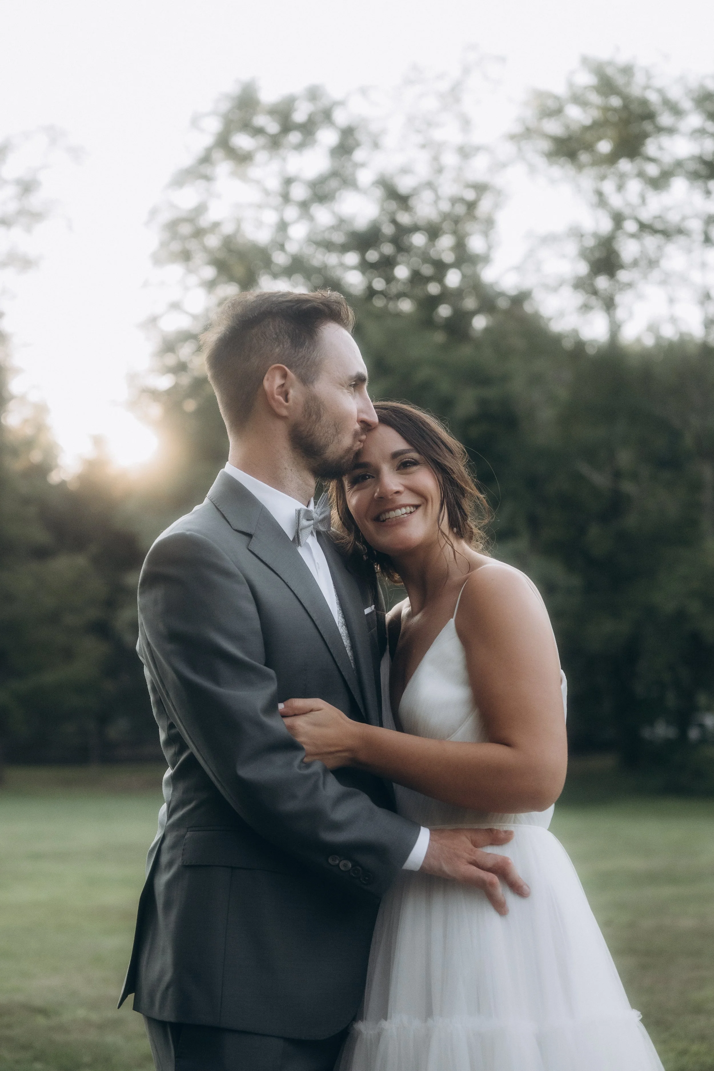 A newlywed couple, a man in a gray suit and woman in a white dress, embrace outdoors during sunset with trees in the background.