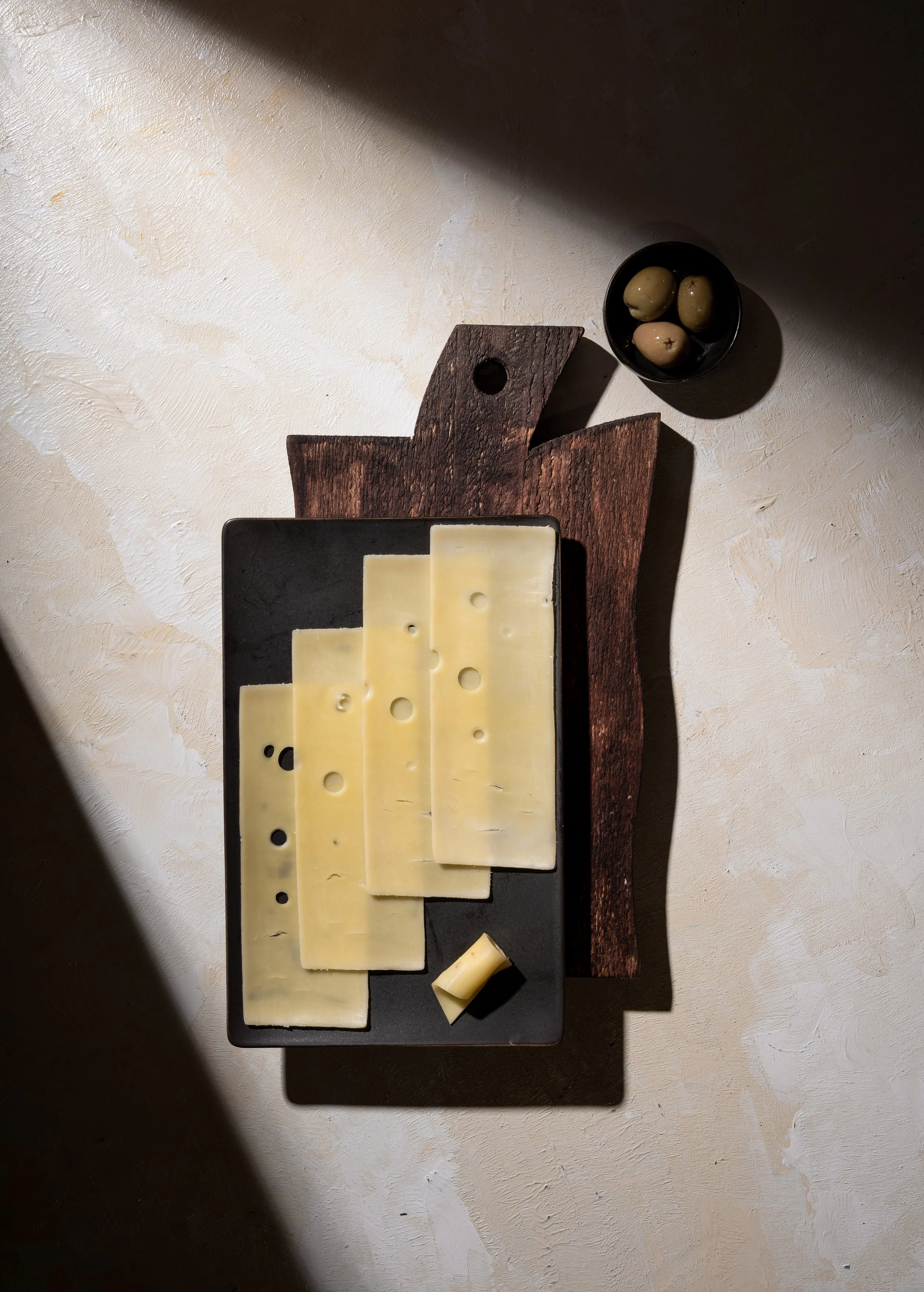A black tray with slices of Swiss cheese with holes, stacked on top of a wooden cutting board, on a light textured surface. A small slice of cheese is in front of the stack. A small black bowl with green olives is in the top right corner.