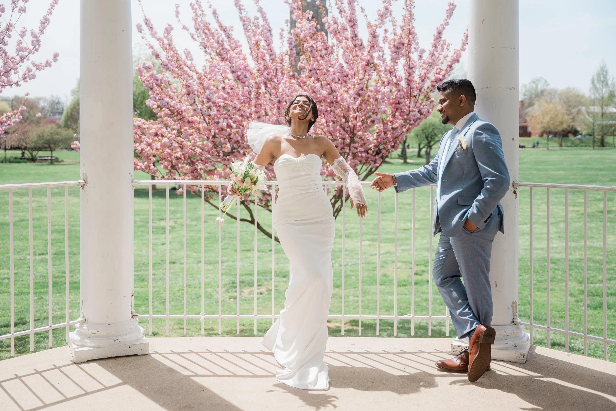 A bride and groom smiling and posing for a photo on a gazebo with blooming pink cherry blossom trees in the background on a sunny day.