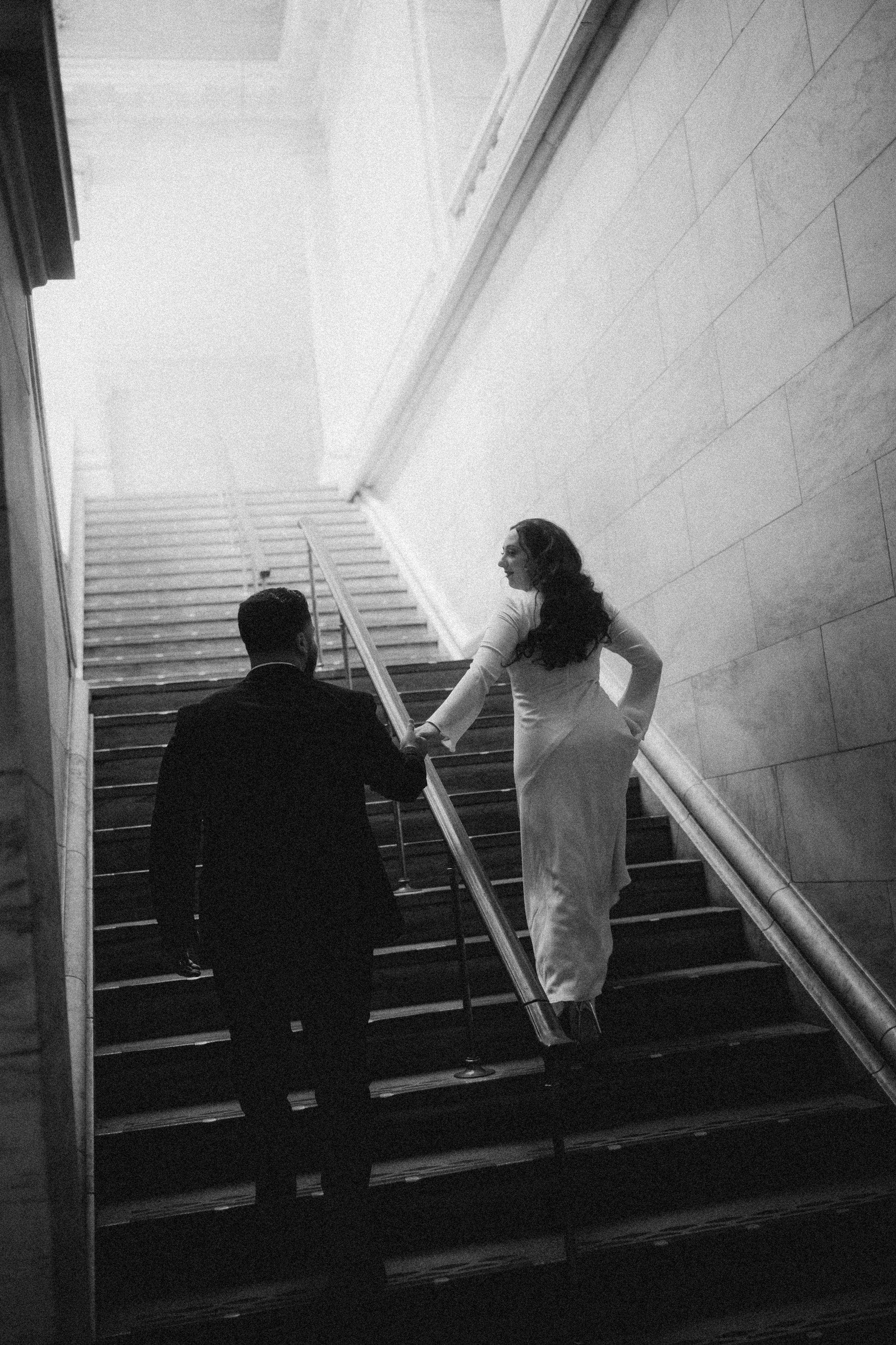 A black-and-white photo of a man dressed in a dark suit holding a woman's hand as they ascend a staircase. The woman wears a long white dress and has long curly hair, looking back over her shoulder.