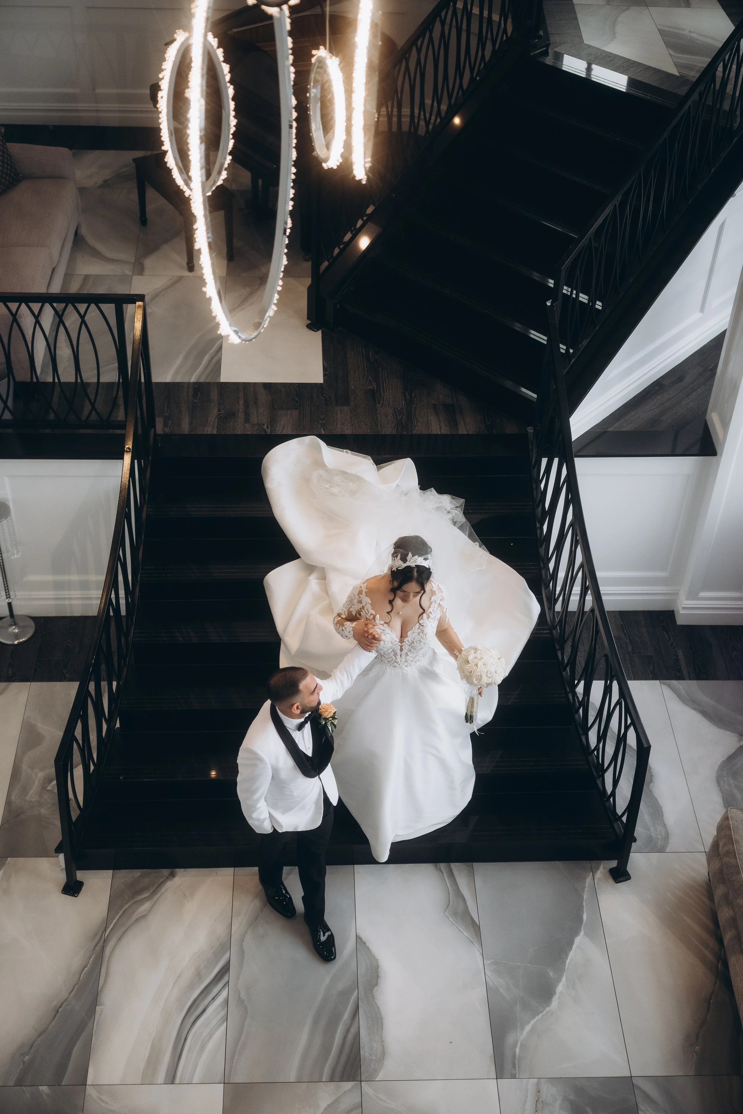 Overhead view of a bride and groom on a staircase, the bride holding a bouquet of white roses, the groom in a tuxedo, as a chandelier with circular light fixtures hangs above them.