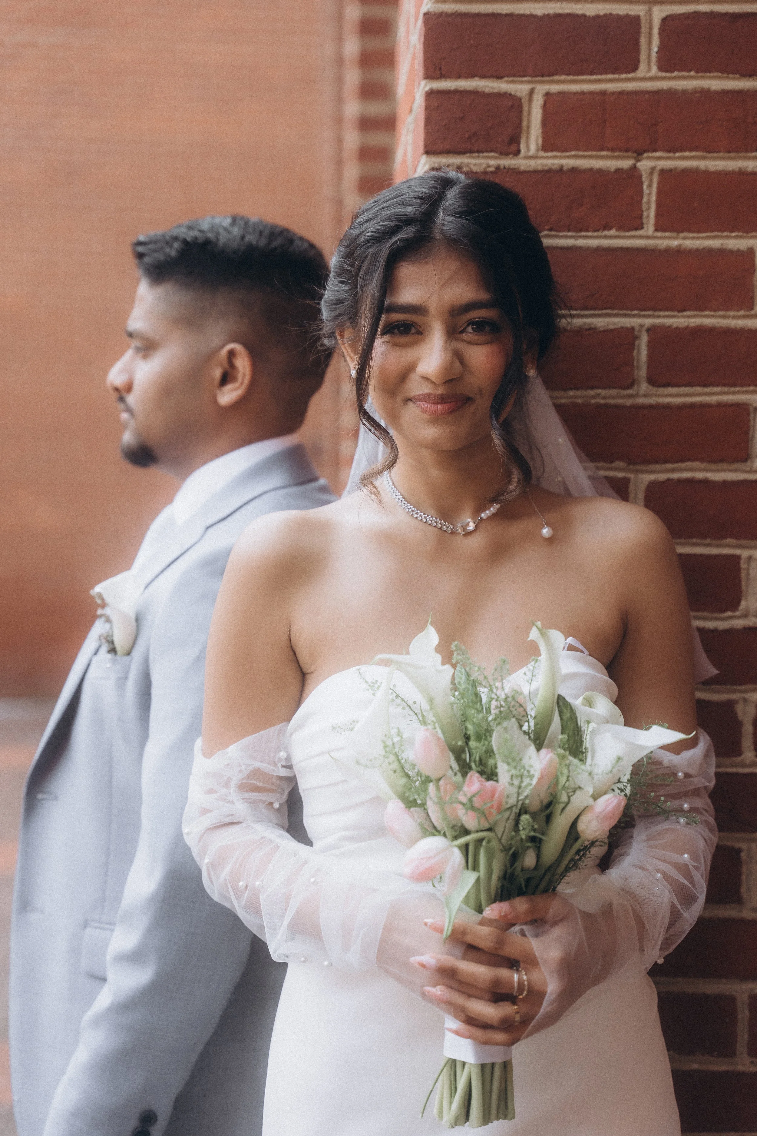 A bride holding a bouquet of white and pink flowers, smiling, standing next to a groom who is facing sideways, against a brick wall.