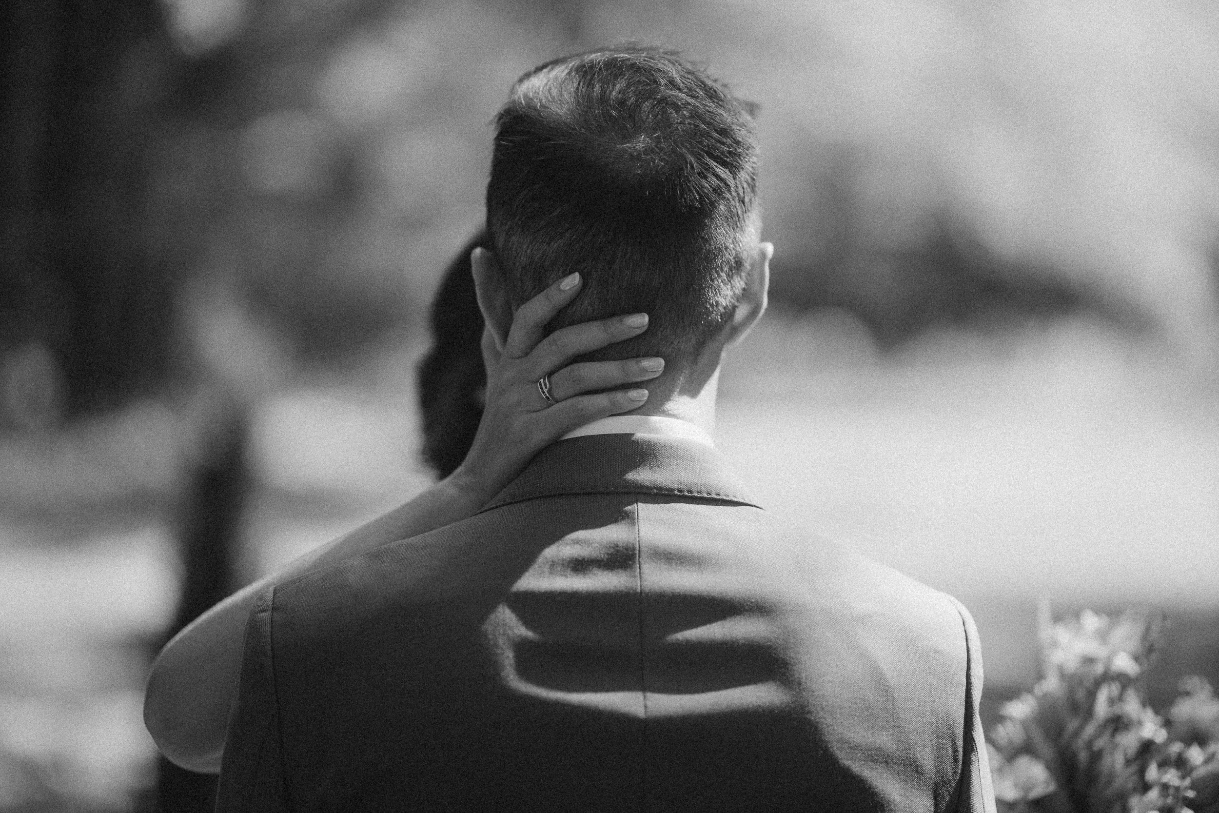 A black and white photo of a couple embracing, with the woman’s hand on the man's neck, showing a ring, as they face away from the camera.
