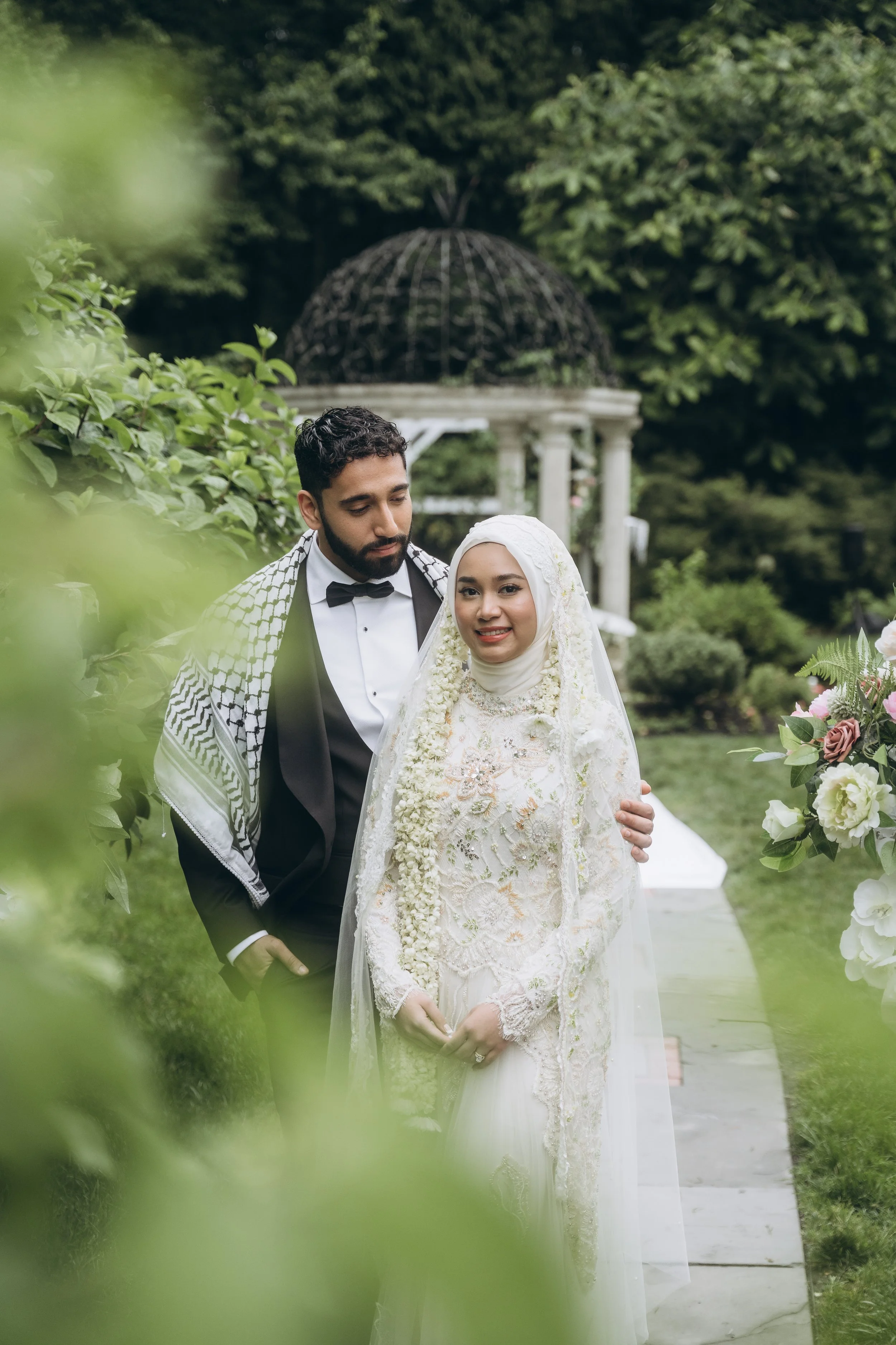 A bride and groom standing together outdoors in a garden, with the groom holding the bride's arm. The bride is wearing a white lace dress and a hijab, and the groom is wearing a black tuxedo with a bow tie. There are green trees and a decorative gaze