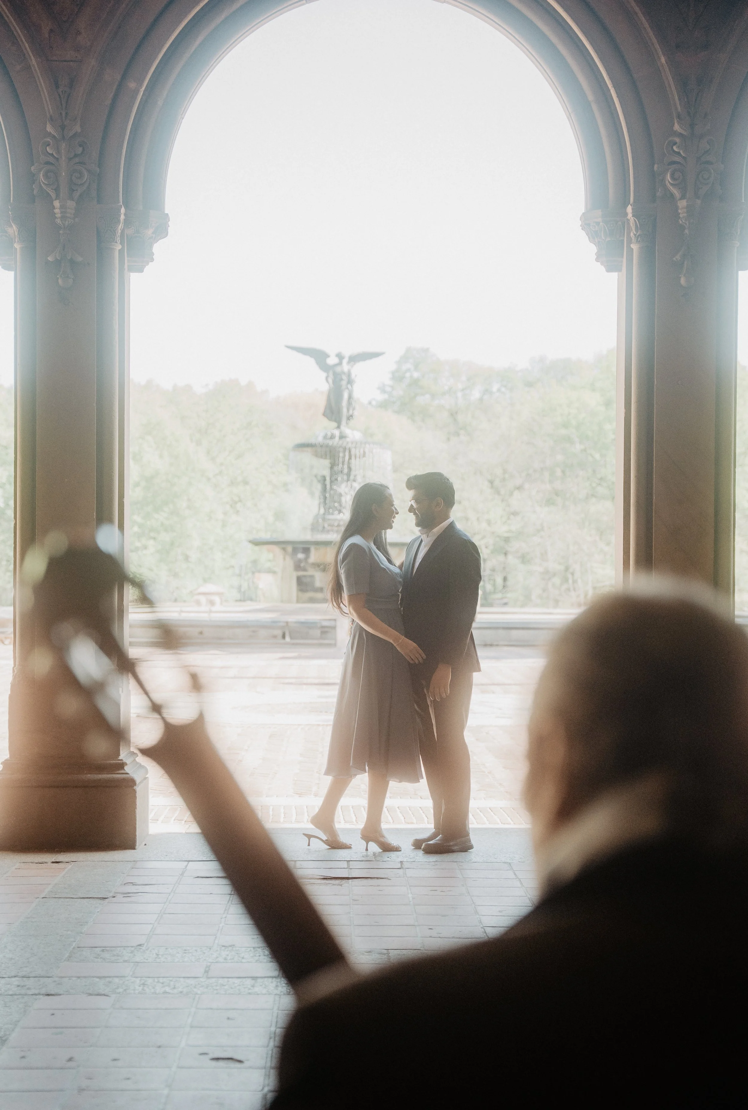 A couple stands close together, dancing and smiling in front of a large open archway with a statue in the background in Central Park NYC, and a person playing a violin in the foreground.