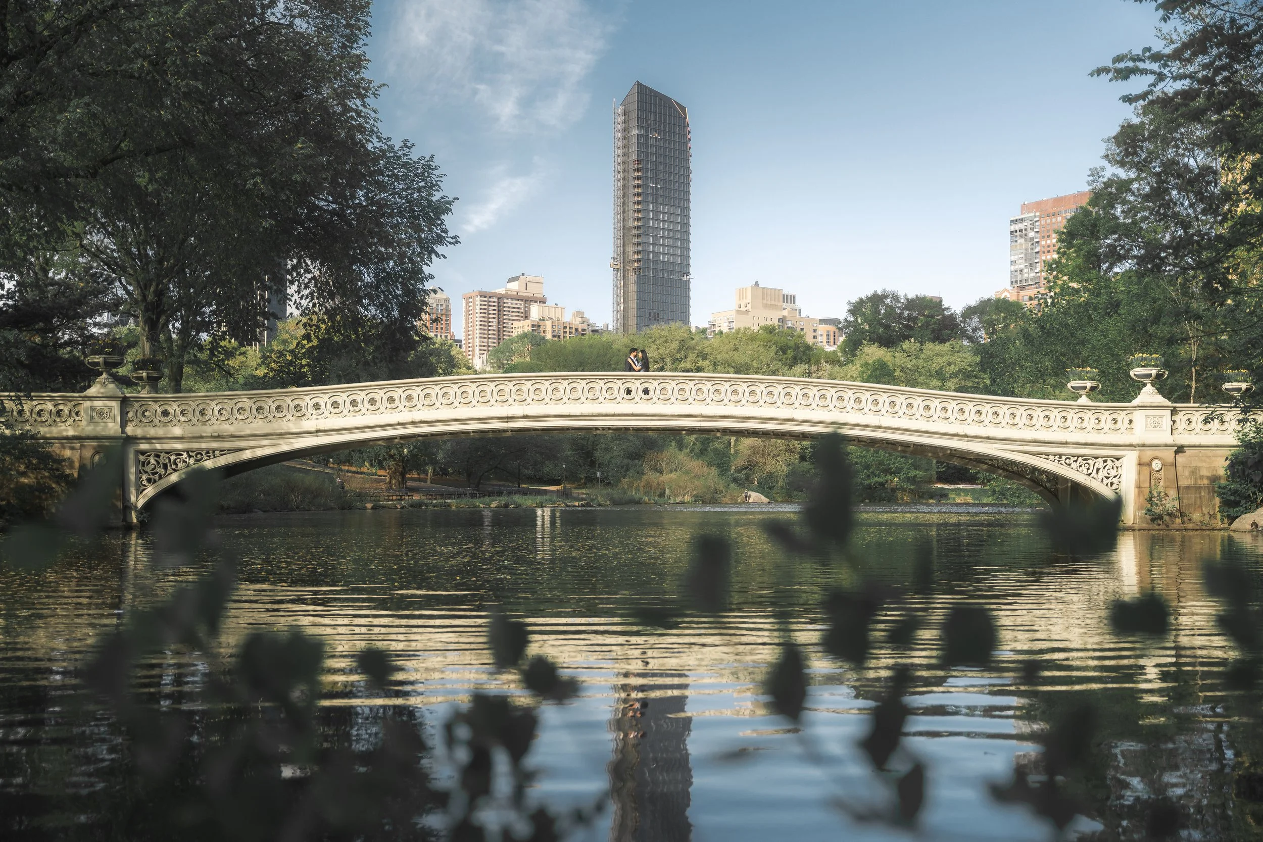 A decorative white bridge (The Bow Bridge)  over a lake in Central Park NYC, with a city skyline and a tall building in the background, and trees surrounding the scene.