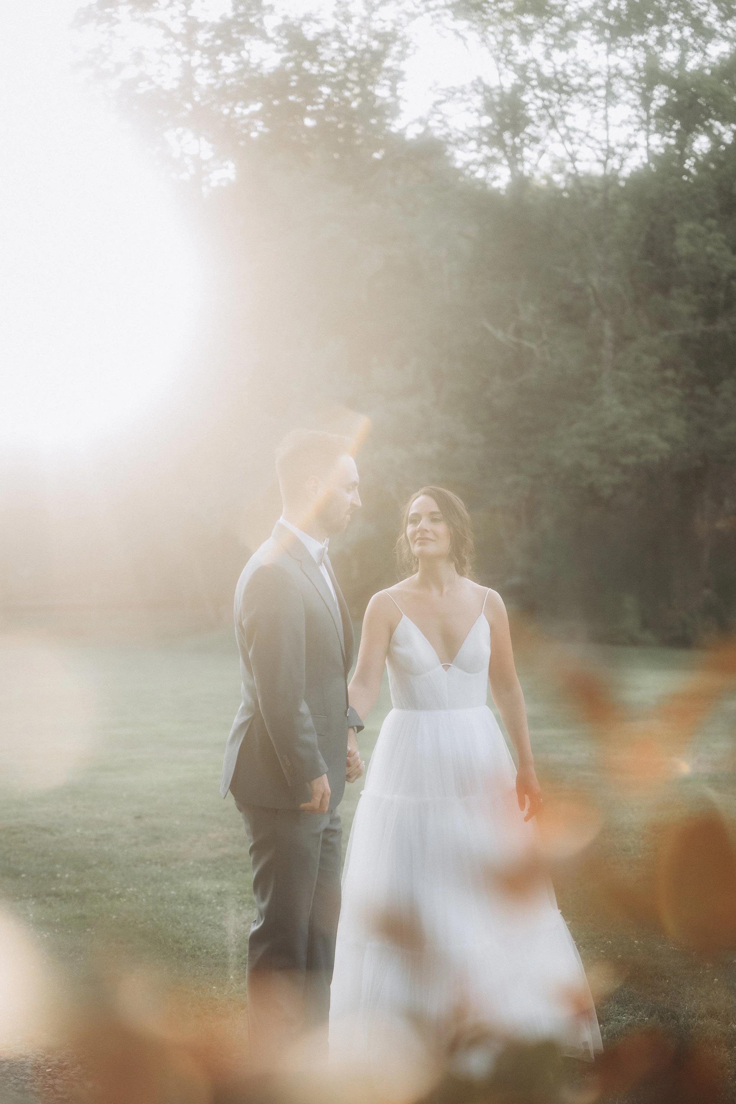 A bride and groom holding hands outdoors during a wedding photoshoot, with trees and greenery in the background, and a hazy light effect.