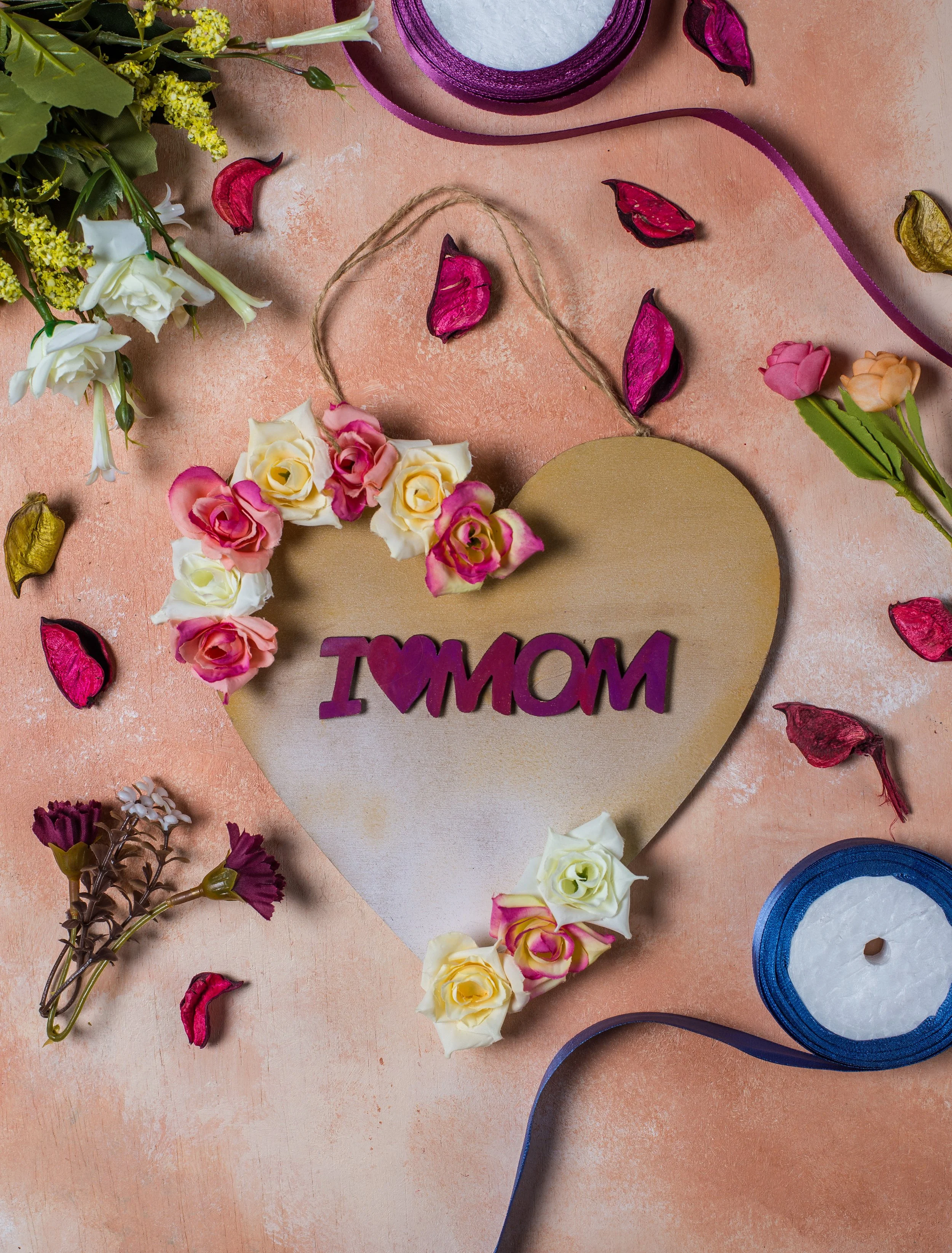 A heartfelt Mother's Day decoration featuring a wooden heart with a purple "I ❤️ MOM" sign, surrounded by small roses, flower petals, and ribbon on a pink textured background.