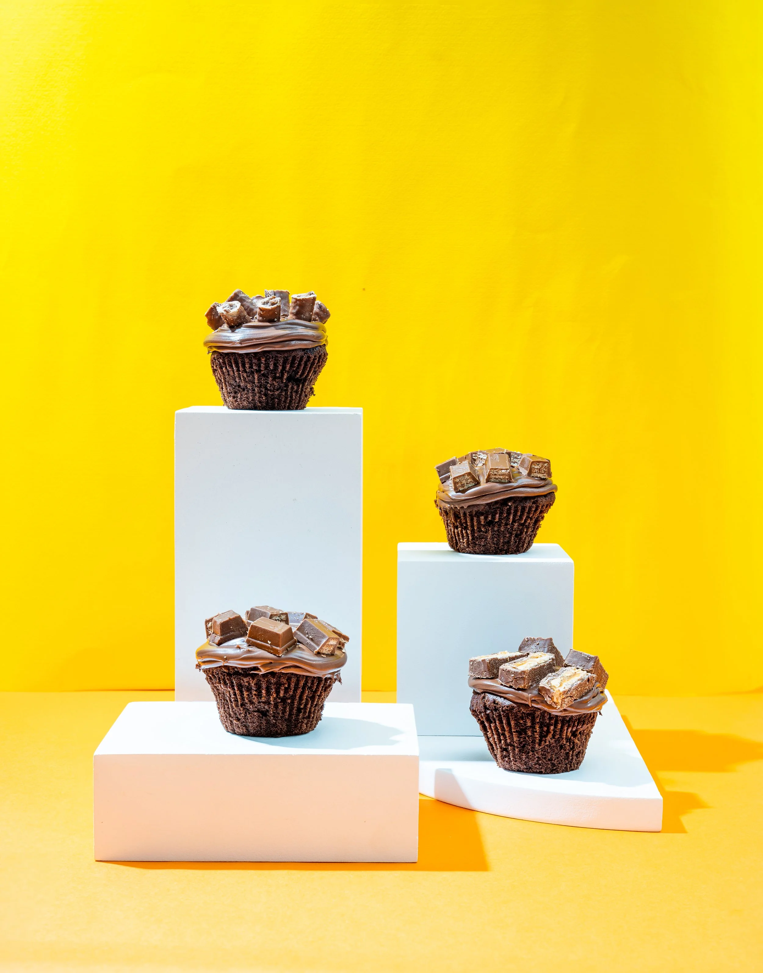 Four chocolate cupcakes with chocolate frosting, topped with various chocolate chunks, displayed on white and yellow podiums against a yellow background.