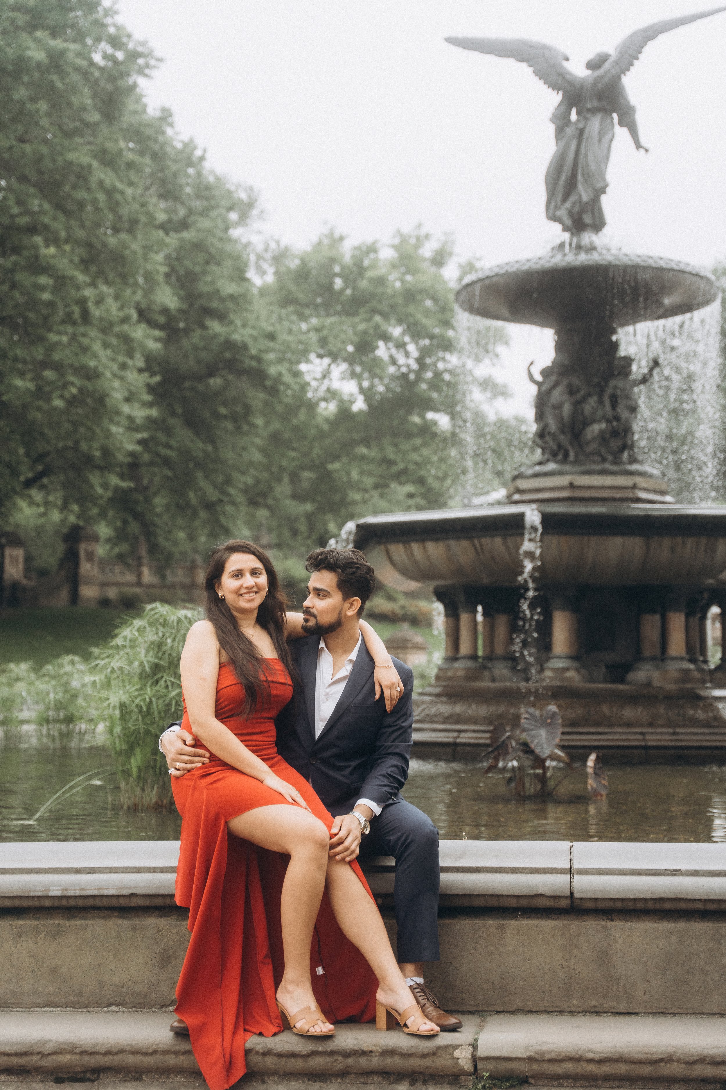 A couple sitting on the edge of a fountain in Central Park NYC, with the woman in a red dress and the man in a suit with an open collar, in a park with trees in the background.