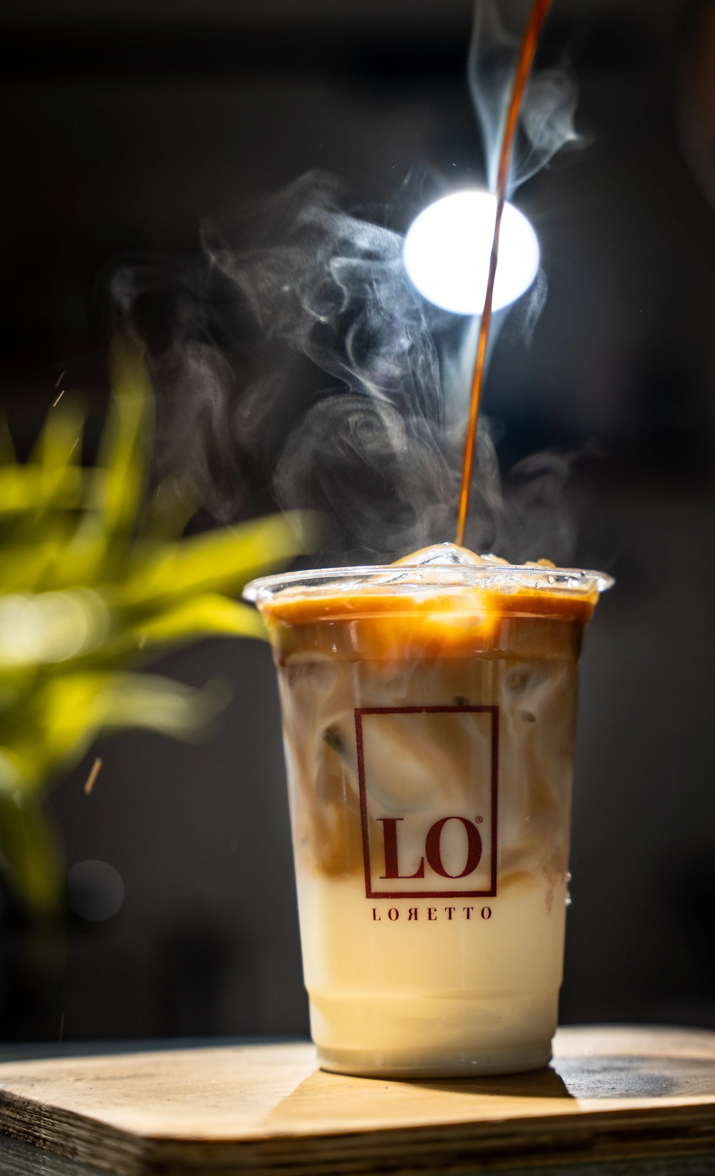A cold coffee drink with swirls of cream in a clear plastic cup labeled 'LO' with some steam rising, placed on a wooden surface.