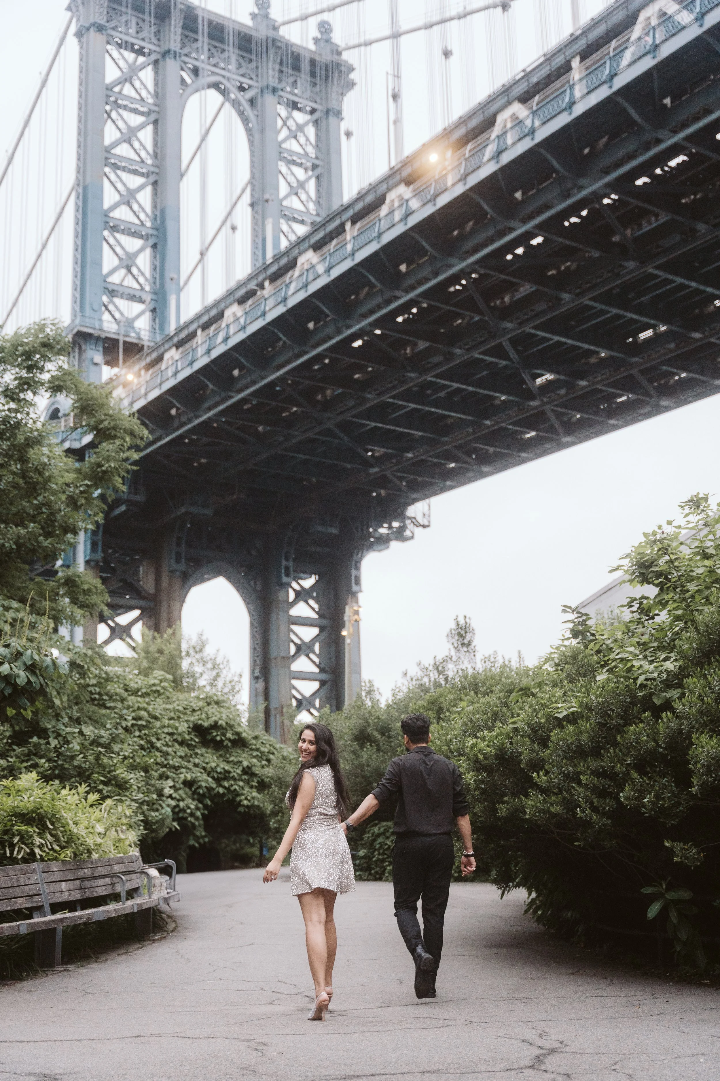 A couple holding hands walking under a bridge in a park in Dumbo Brooklyn, with the woman turning back and smiling at the camera.