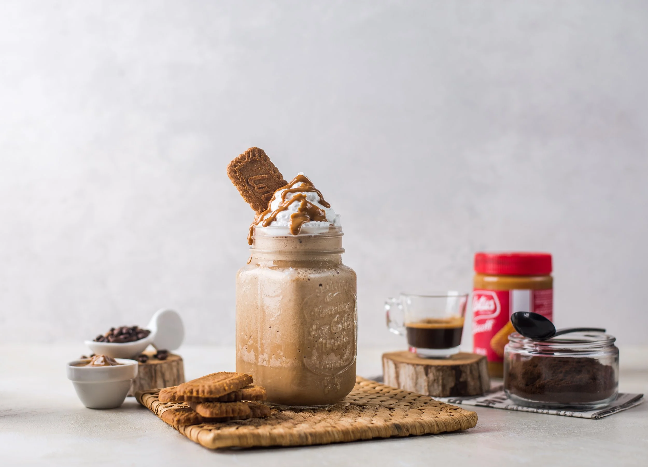 A mason jar filled with a creamy coffee beverage topped with whipped cream, caramel sauce, a cookie, and a biscuit. Surrounding the jar are small bowls and jars of coffee beans, ground coffee, coffee powder, and instant coffee, on a woven mat and a w
