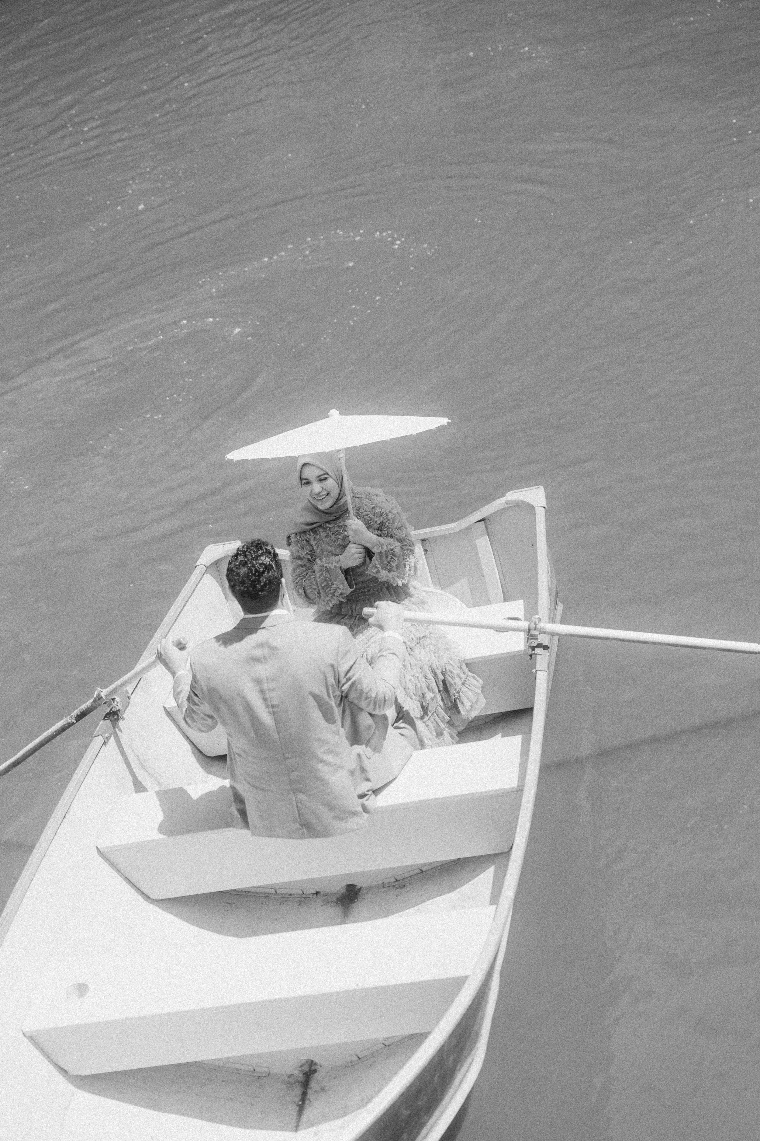 A black and white photo of a woman in a dress and hijab sitting in a rowboat holding an umbrella, with a man in a suit sitting in front of her. They are on lake in Central Park NYC, seen from above.