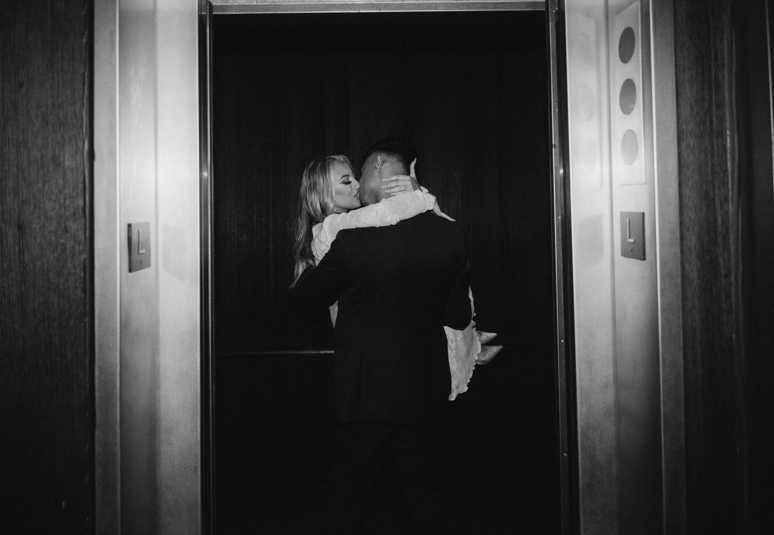 A man in a suit is holding a woman in a white dress inside an elevator, as she kisses him with her arms around his neck.
