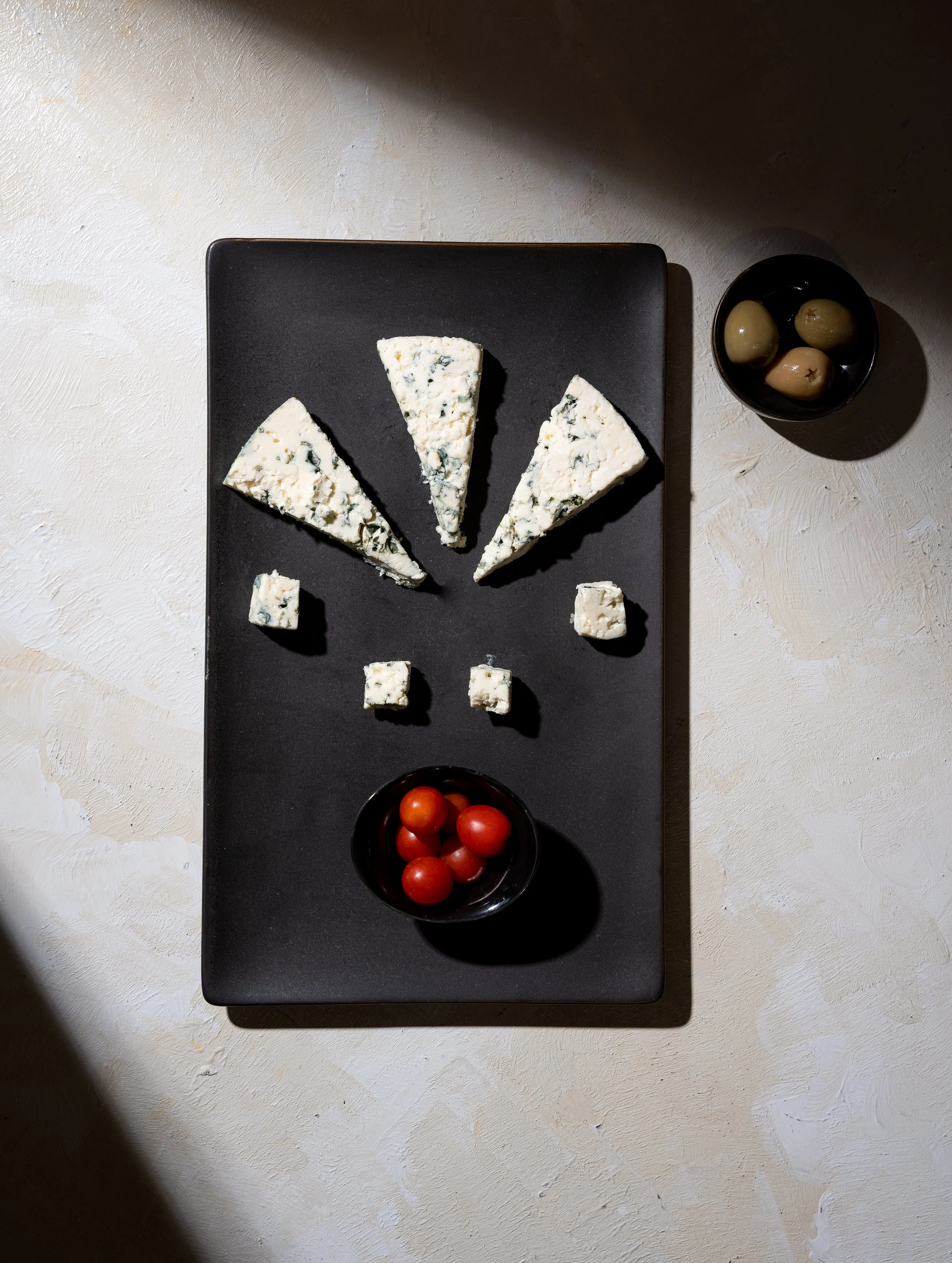 A black rectangular platter with four slices of blue cheese, four cubes of blue cheese, and two small black bowls containing green and yellow olives and cherry tomatoes, arranged on a white textured surface with light shadows.