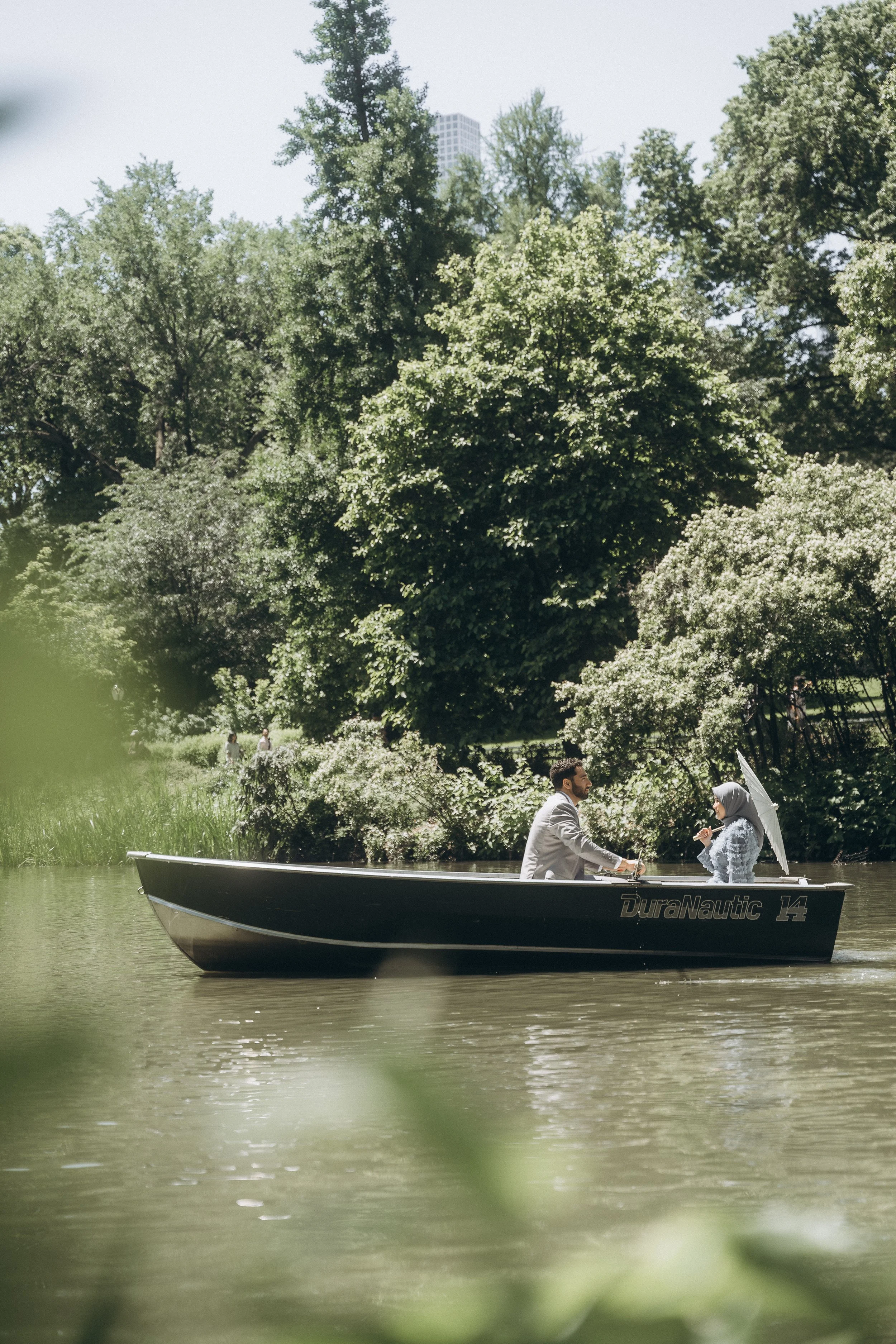 A man and woman in formal attire sitting in a boat on a lake in Central Park NYC, surrounded by greenery and trees.