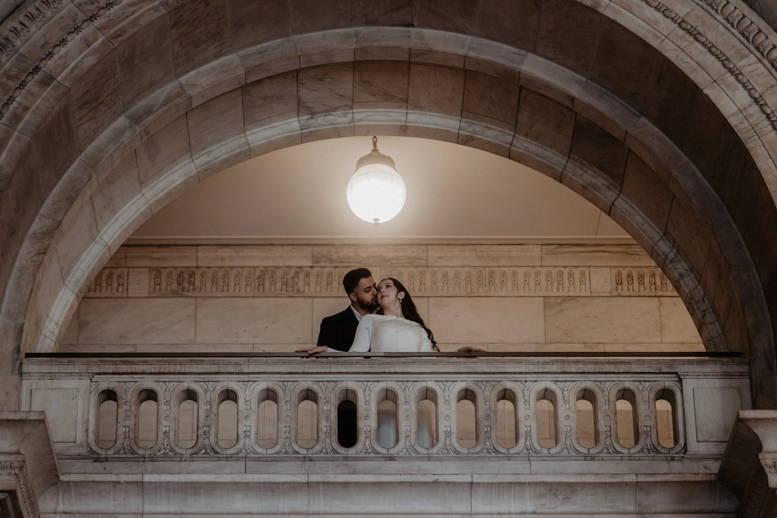 A couple sharing a kiss on a balcony inside a grand building with marble architecture.