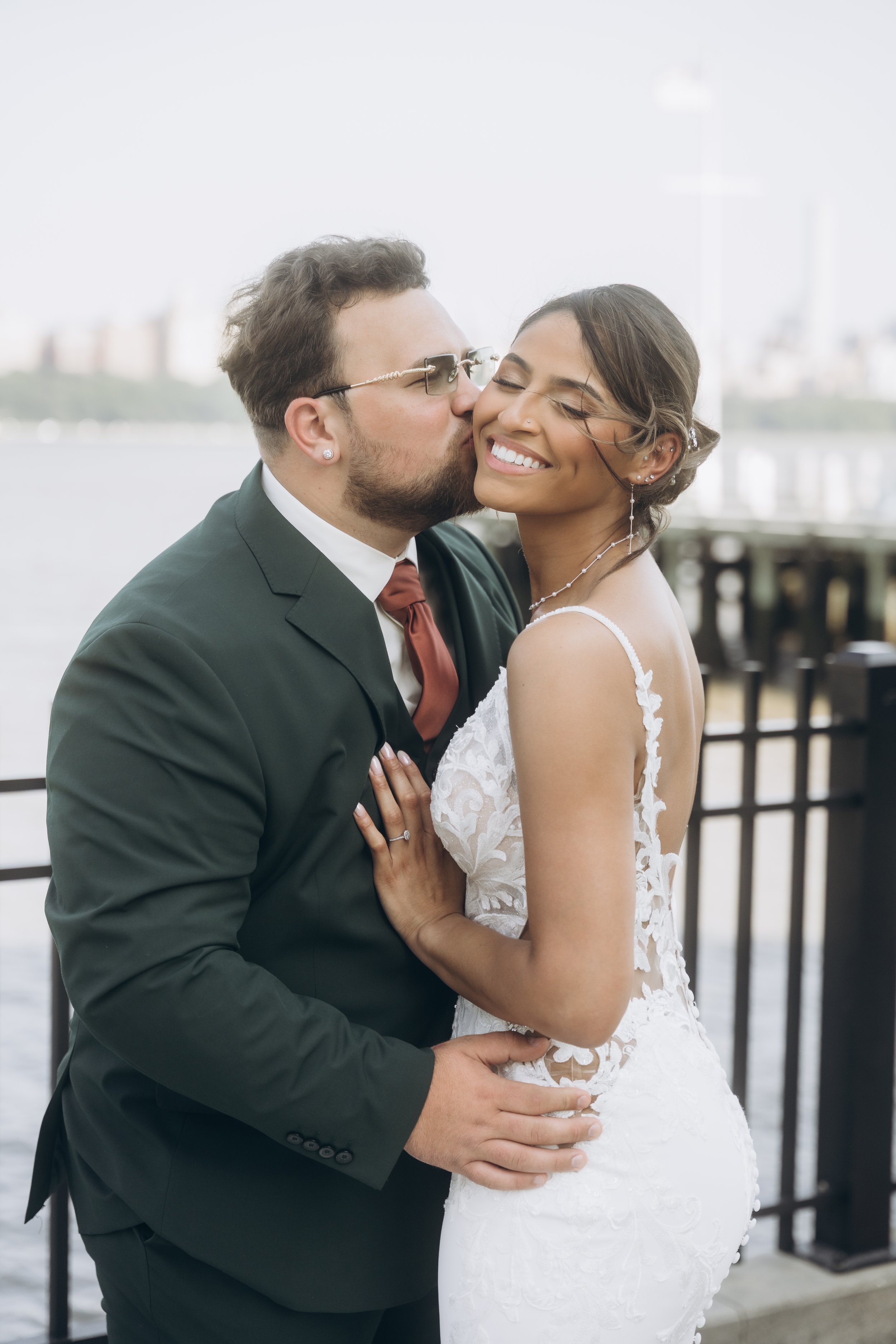 A newlywed couple embracing by a waterfront, with the groom kissing the bride's cheek, both smiling.