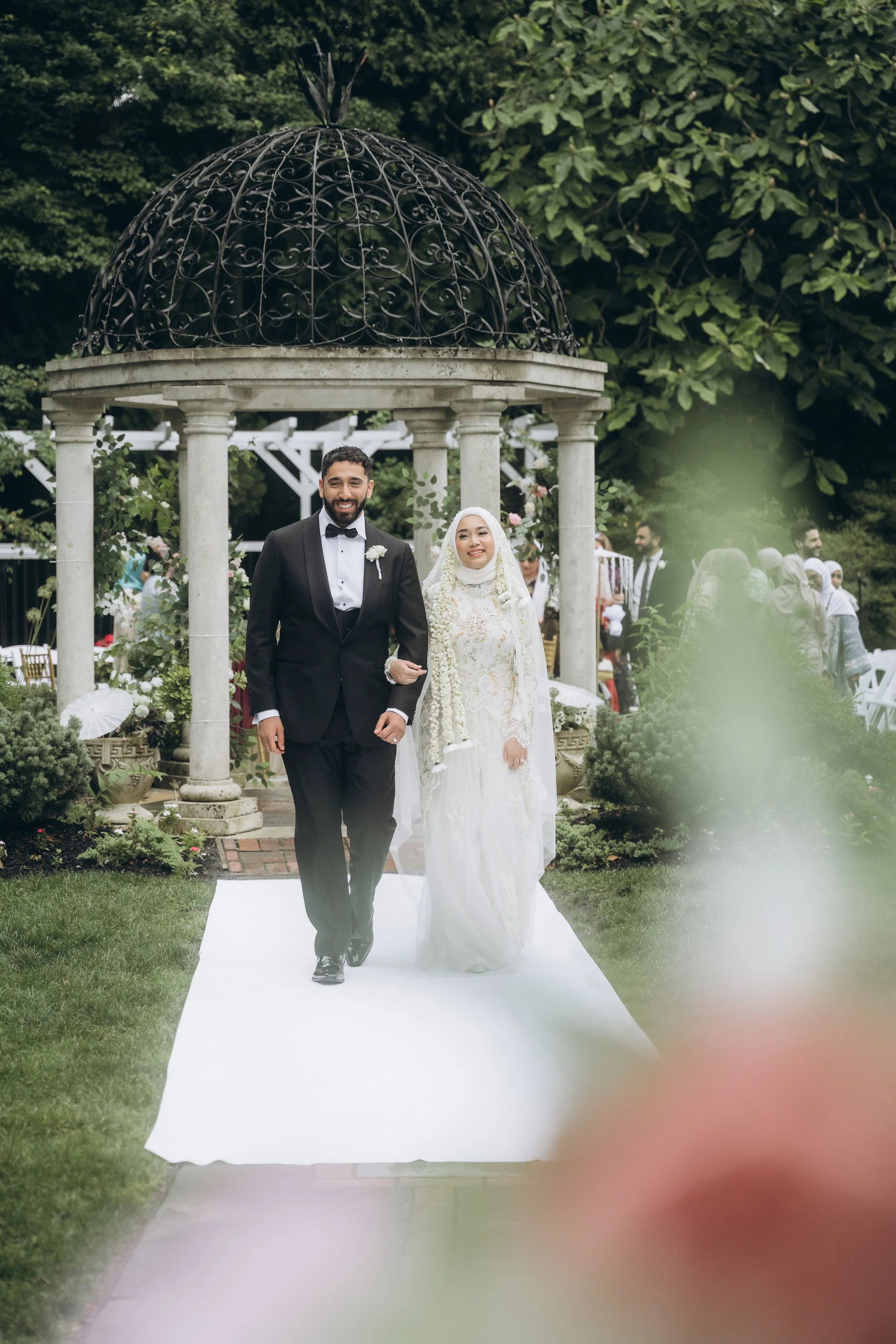 A newlywed couple walking down an outdoor aisle, with the groom in a tuxedo and the bride in a white wedding dress and hijab, under a decorative pavilion, surrounded by greenery and wedding guests.