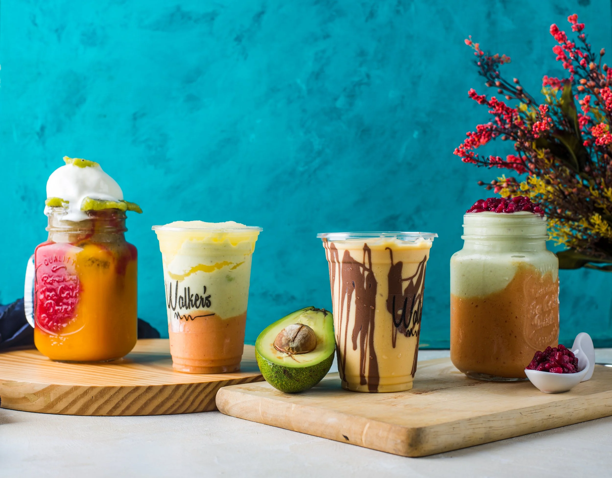 Four jars and cups of colorful smoothies with various toppings on wooden boards, a halved avocado, a spoonful of berries, and a bouquet of colorful flowers against a teal background.