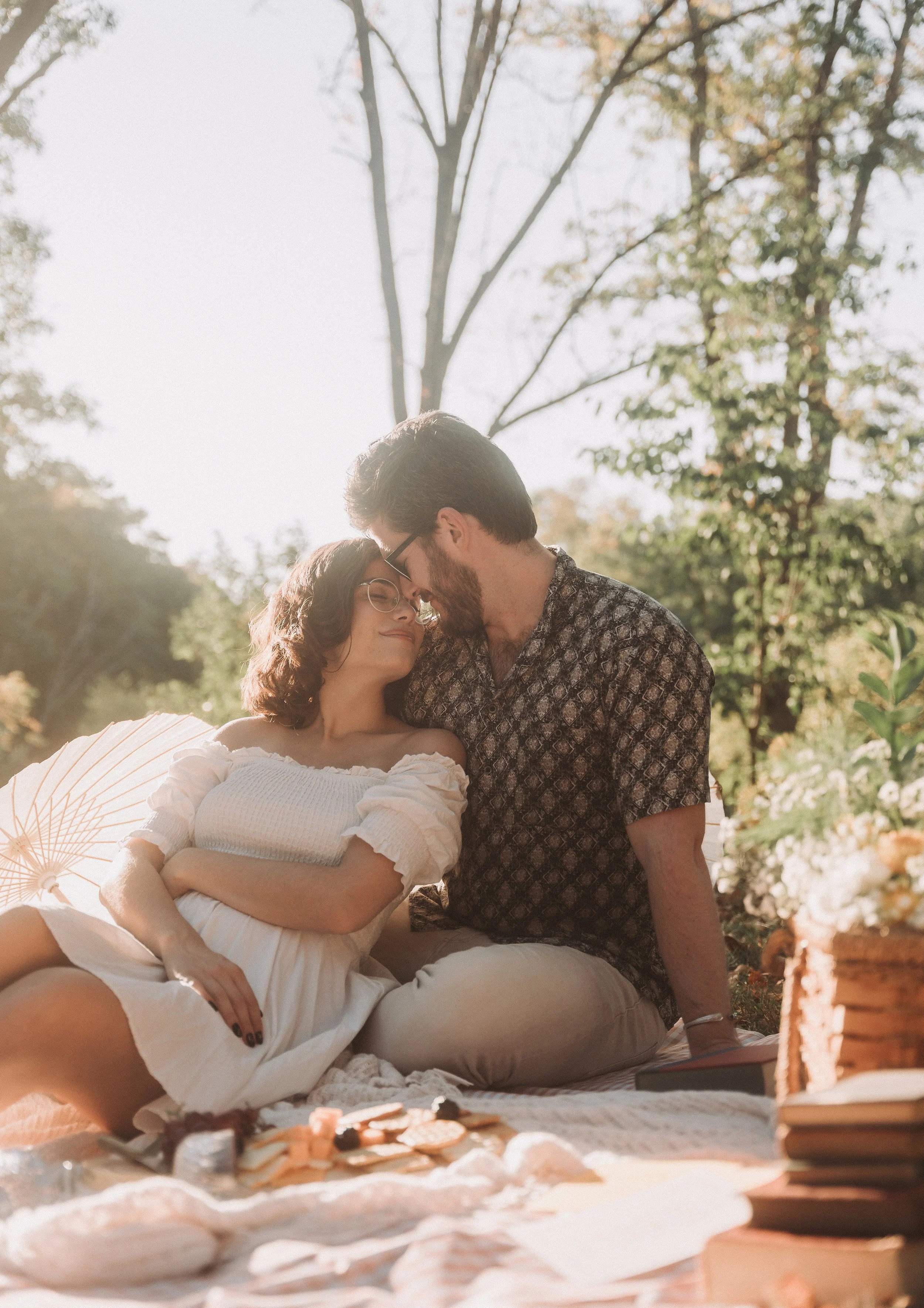 A couple sharing an intimate moment during a picnic outdoors in a forested area on a sunny day.