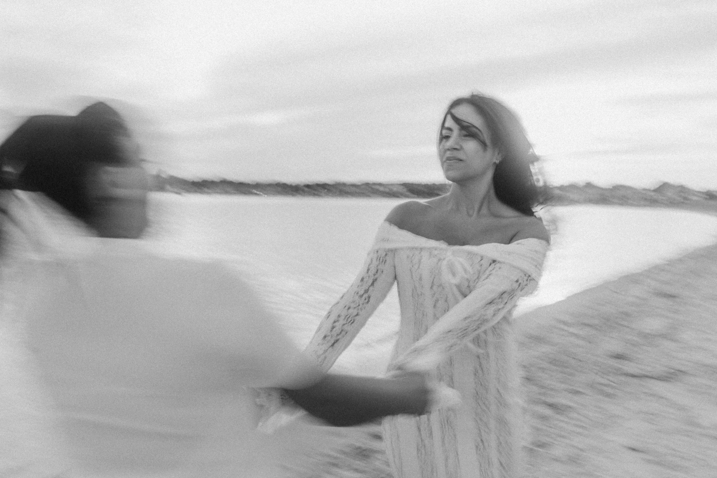 Black and white photo of two women on a beach, holding hands and facing each other, one looking at the other with a serious expression, with the ocean in the background.