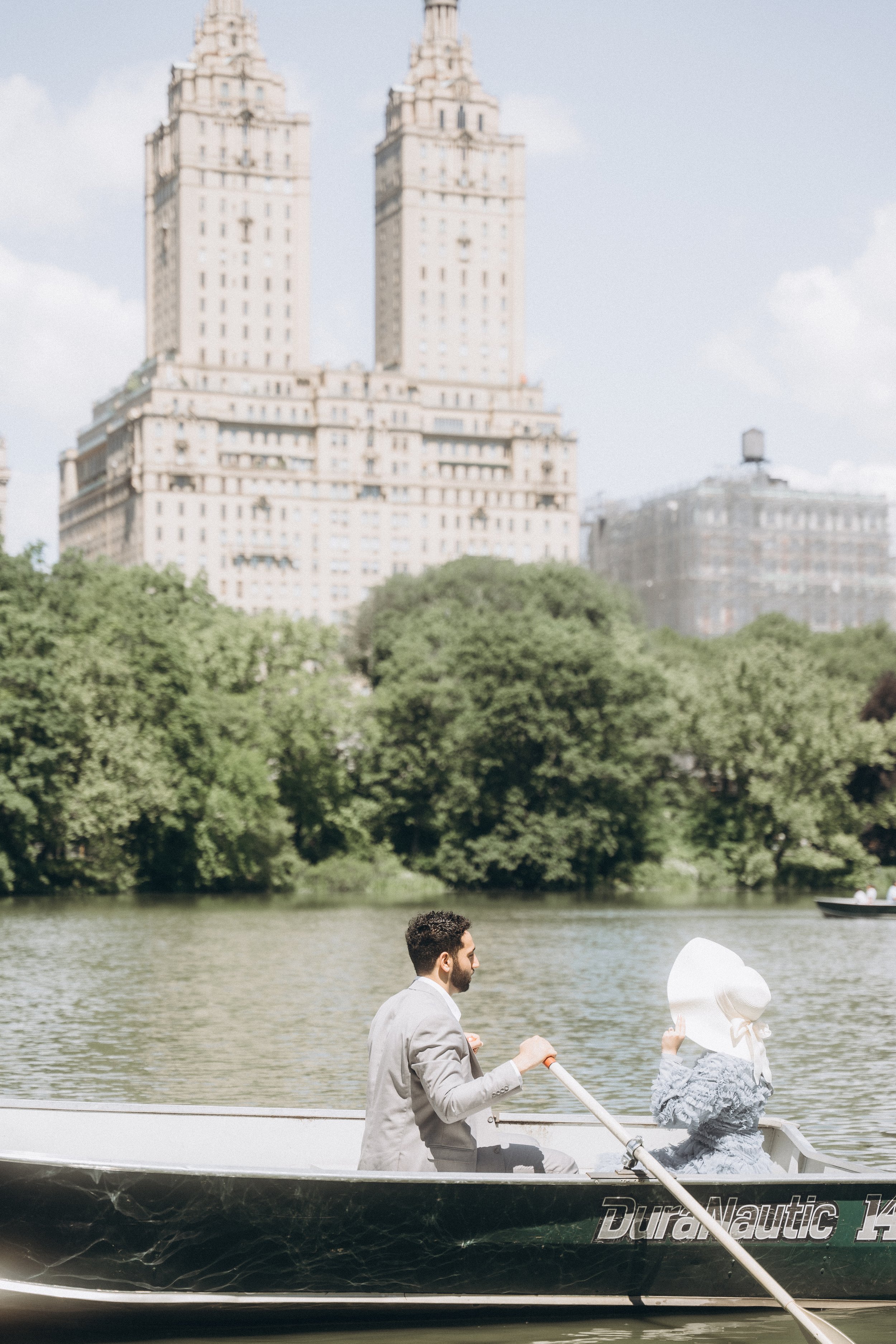 A man and a woman in a boat on a lake in Central Park (NYC) with green trees and tall buildings in the background.