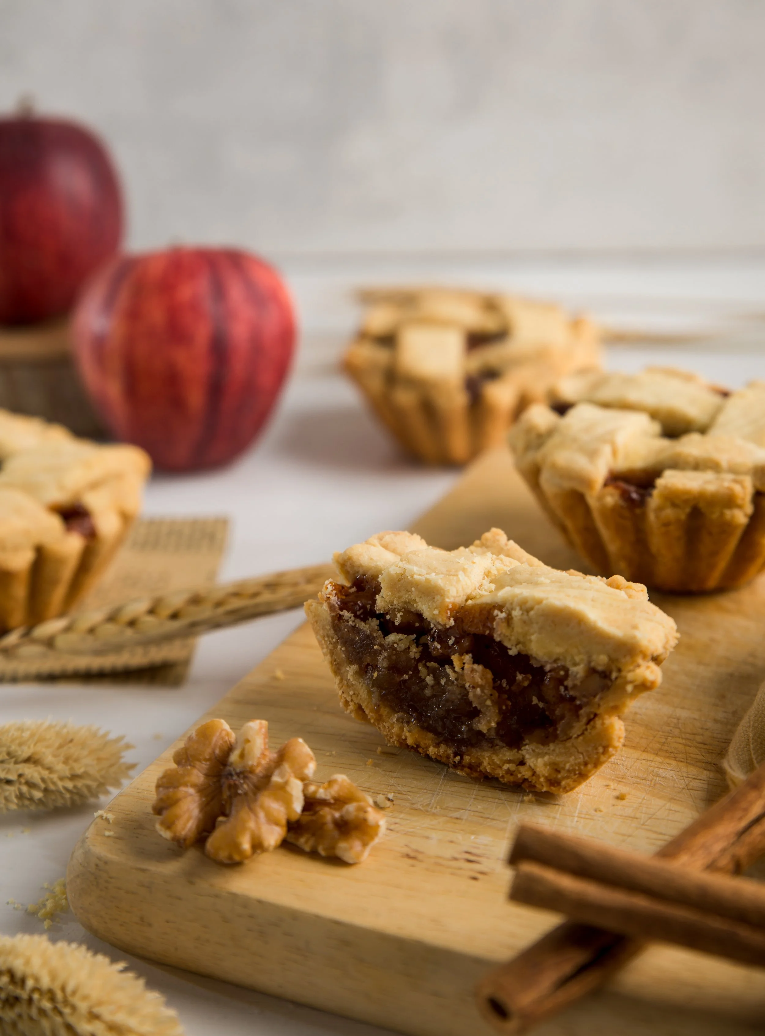 Slices of apple pie on a wooden cutting board with whole apples, cinnamon sticks, and walnuts in the background on a white surface.