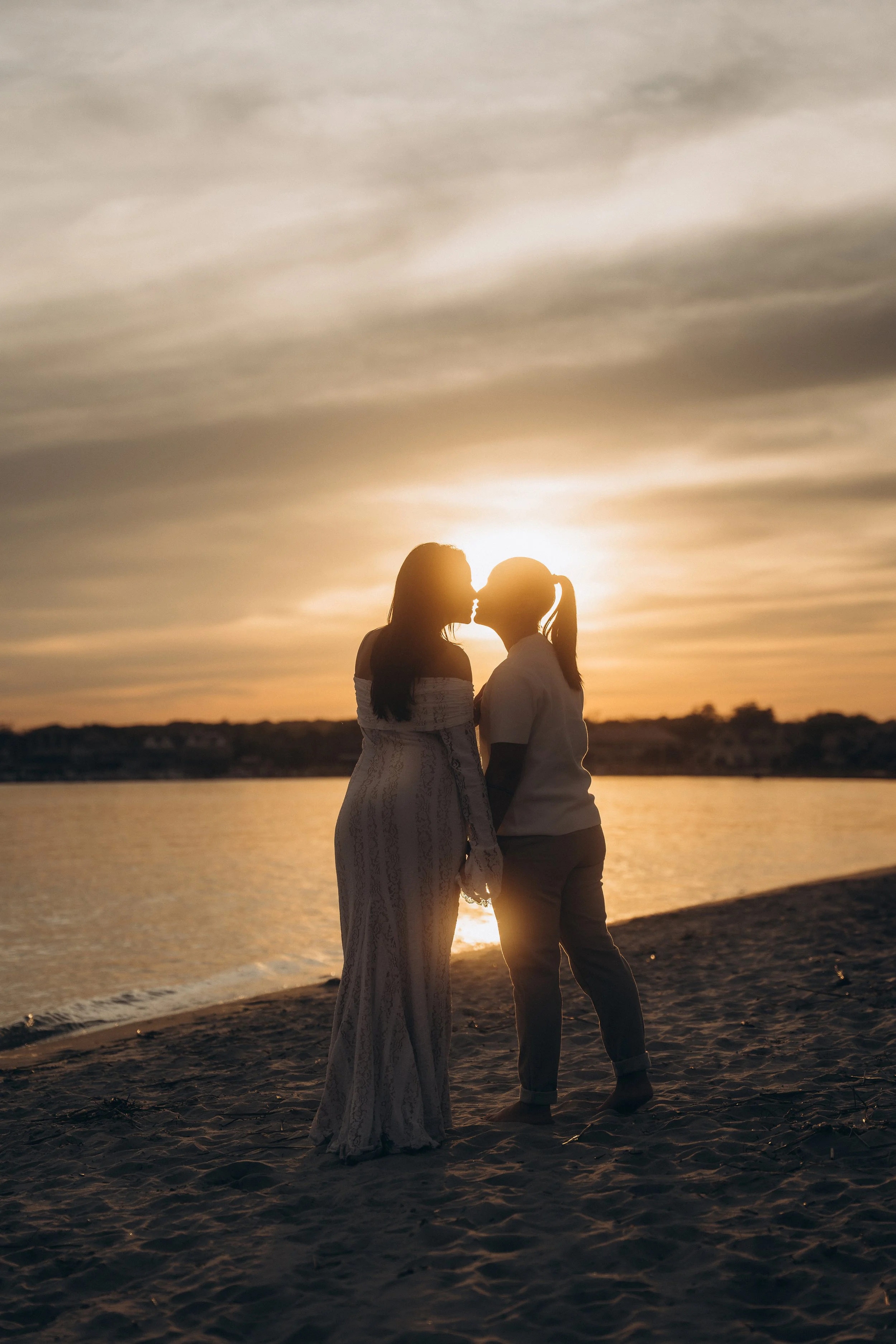 A silhouette of two women standing close on a beach at sunset, about to kiss, with the sun behind them and water and distant land in the background.