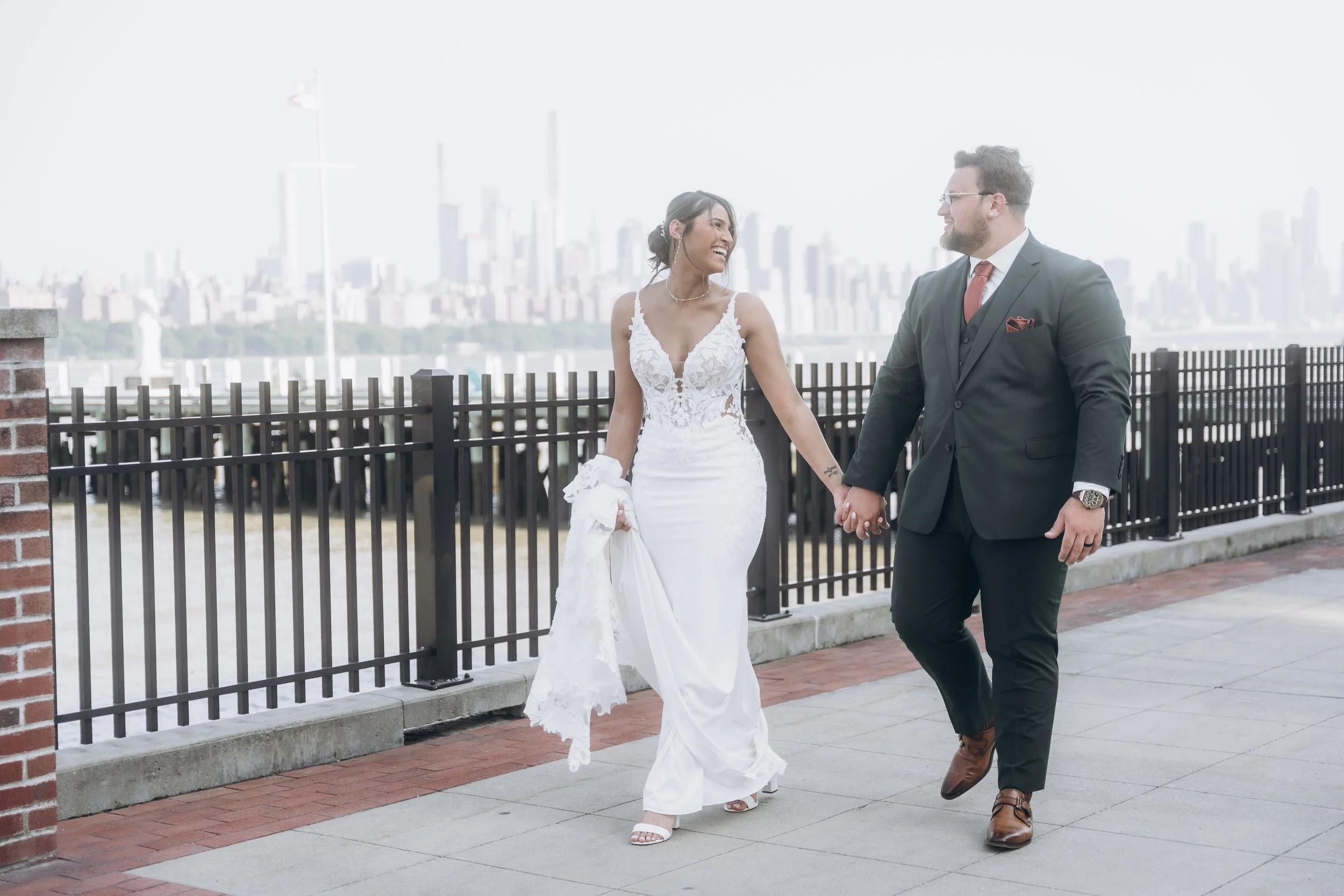 A newlywed couple holding hands and walking outdoors on a paved walkway with a city skyline in the background.