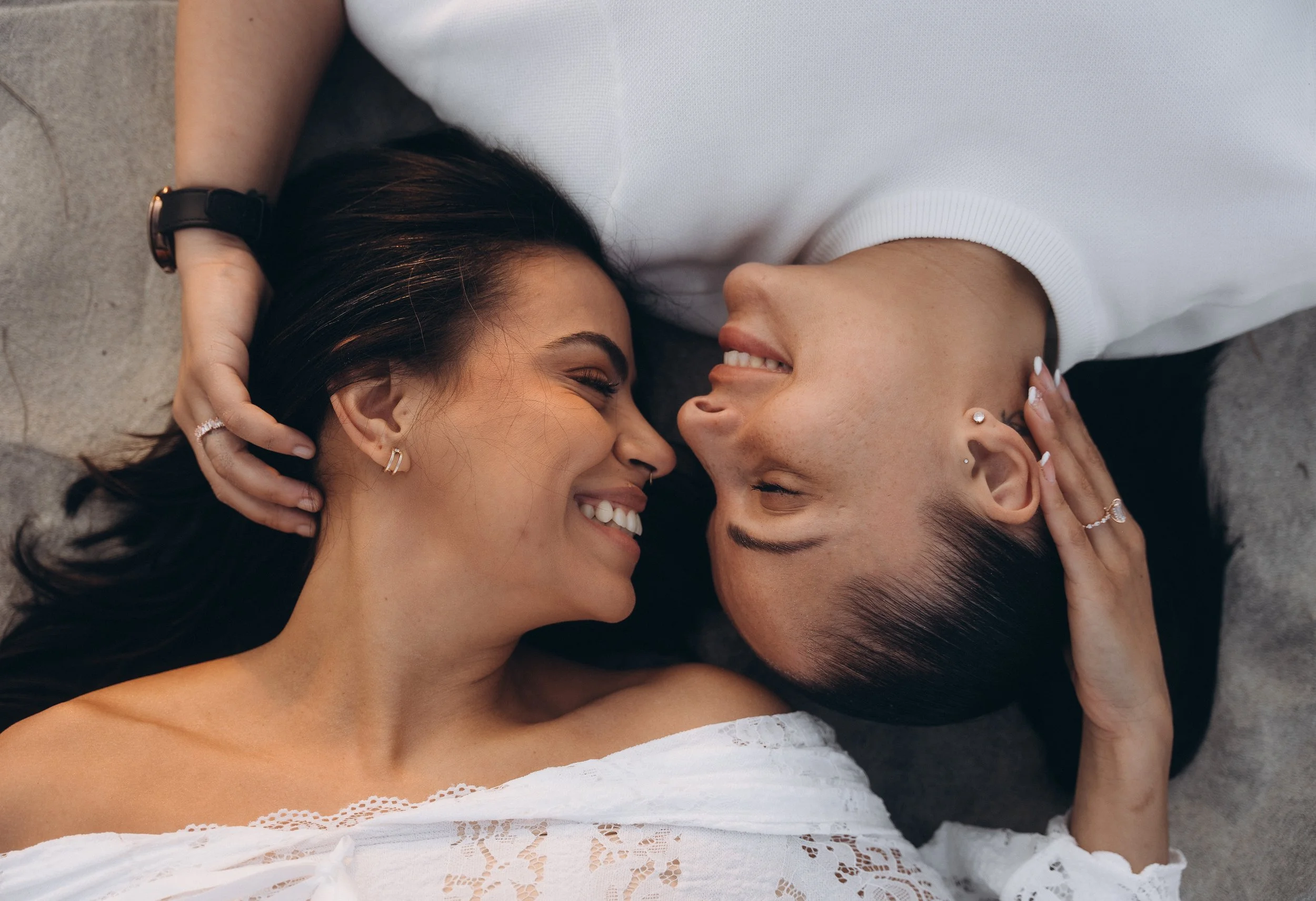 Two women are lying on the floor with their heads touching, smiling at each other. One woman has dark hair and a white top, the other has short hair, piercings and is wearing a white shirt. They are holding each other's heads and appear happy and clo