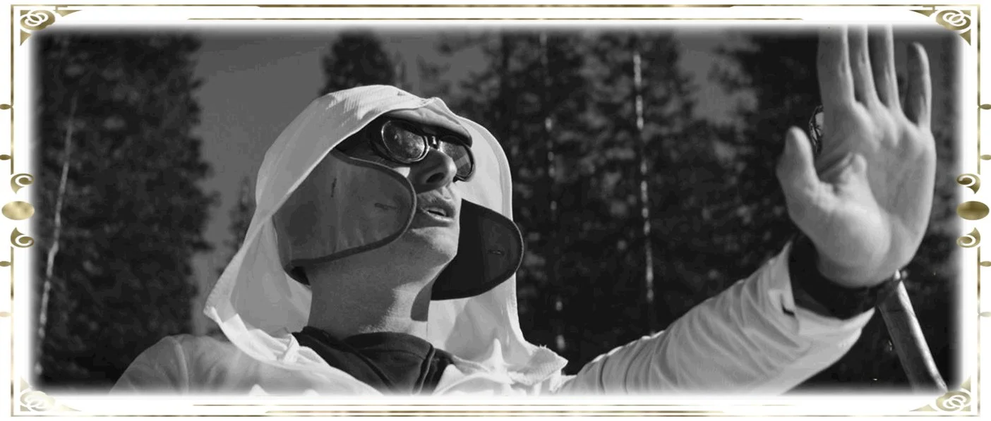 A woman wearing protective gear, including a wide-brimmed hat, goggles, and a disposable suit, is outdoors with trees in the background. She is raising her right hand with her palm facing outward.