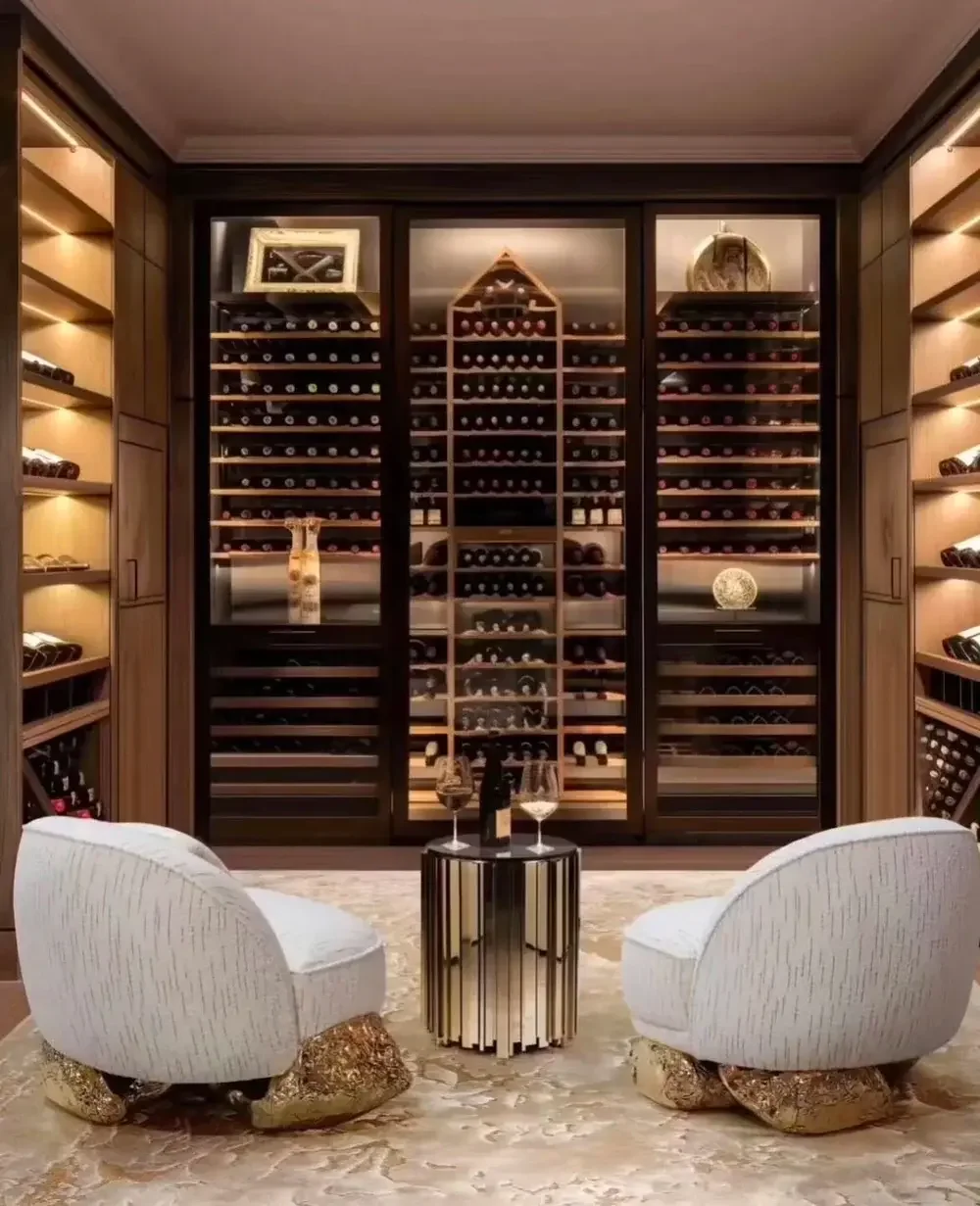 A luxurious wine cellar with wooden shelves filled with wine bottles, a central glass door wine storage, two modern white chairs with gold rock bases, and a small round black table with two wine glasses and a bottle, set on a marble floor.