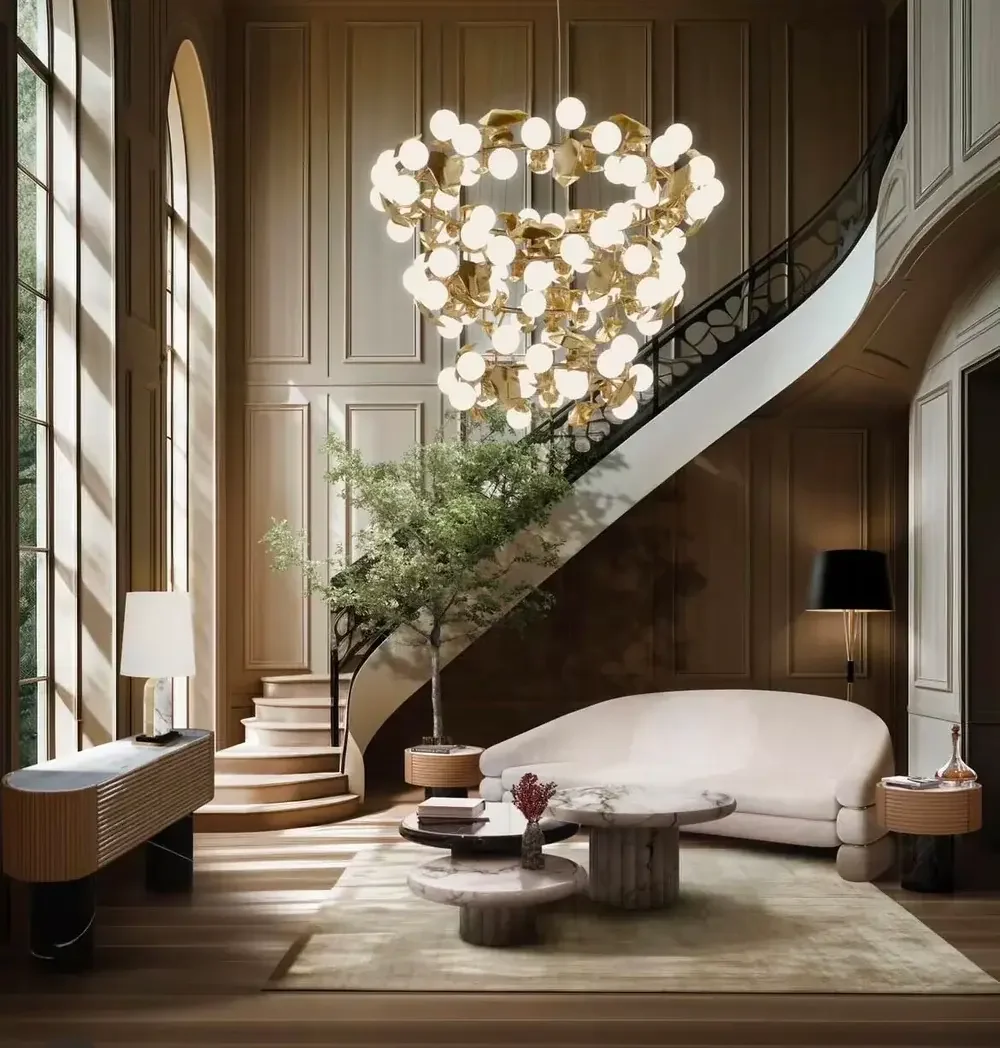 Living room with a grand chandelier, large windows, a white curved sofa, marble coffee tables, a potted tree, and wooden paneling on the walls.