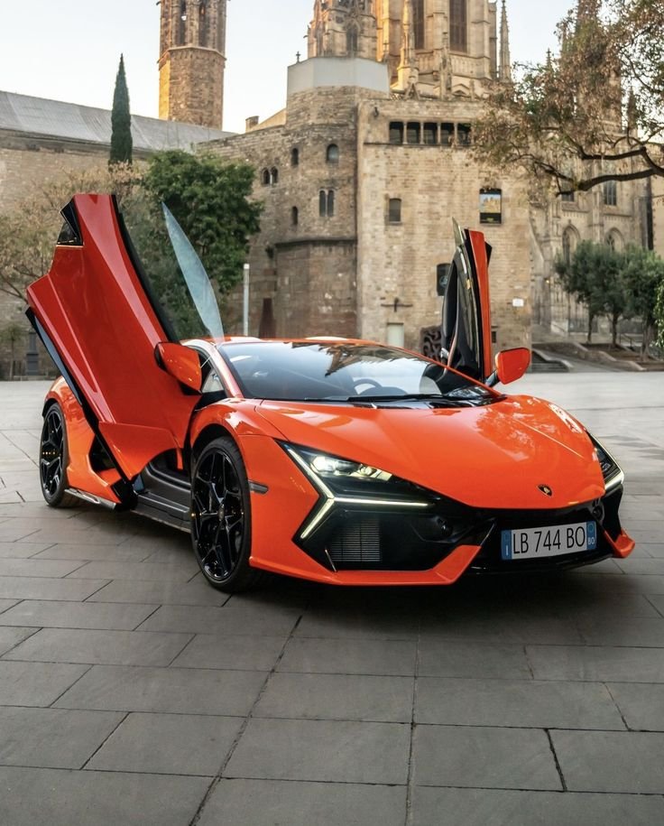 A bright orange Lamborghini sports car with its iconic upward-opening doors open, parked on a stone-paved area in front of a historic stone building with towers and small windows, surrounded by some trees.