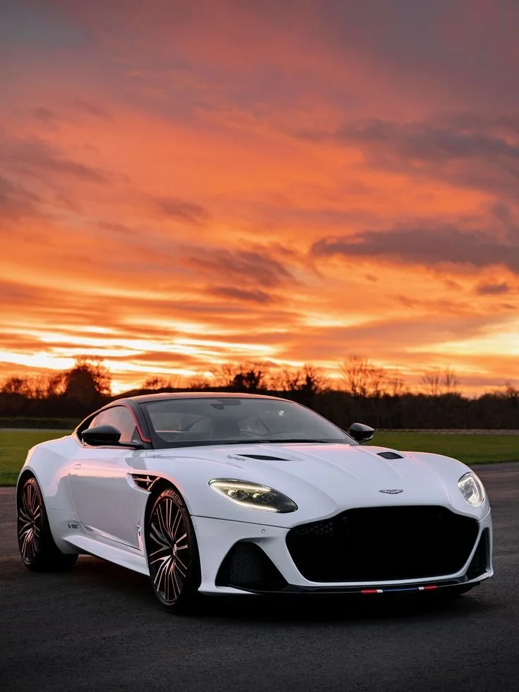 A white sports car parked on a road at sunset with a colorful sky and trees in the background.