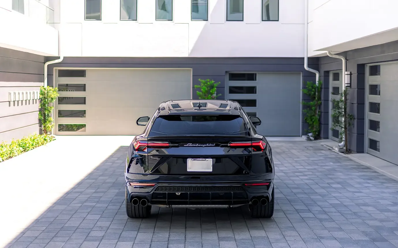 Black Lamborghini Urus parked in a modern driveway with light-colored paving stones, surrounded by a white and gray building with several small trees.