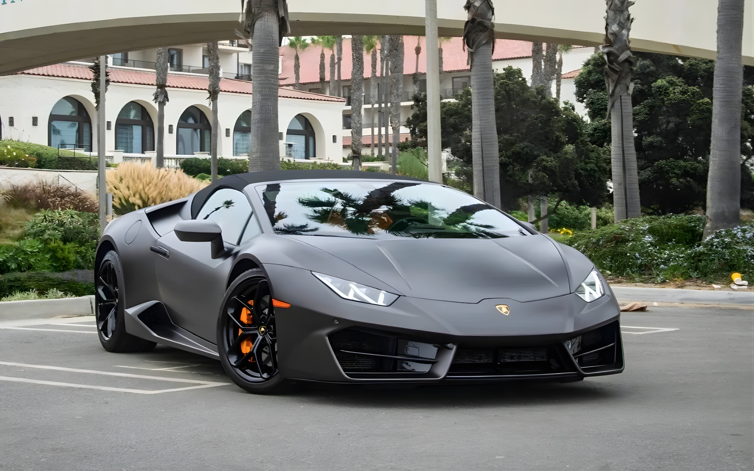 Gray Lamborghini sports car parked in a parking lot with palm trees and buildings in the background.