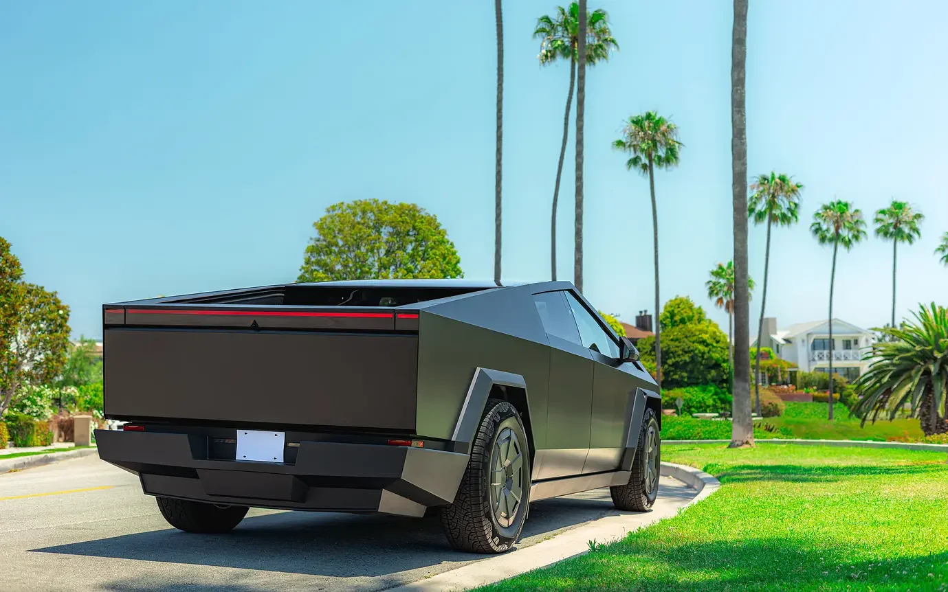 A Tesla Cybertruck Cyberbeast parked on a sunny street lined with palm trees and greenery, showcasing its angular design and matte black finish.