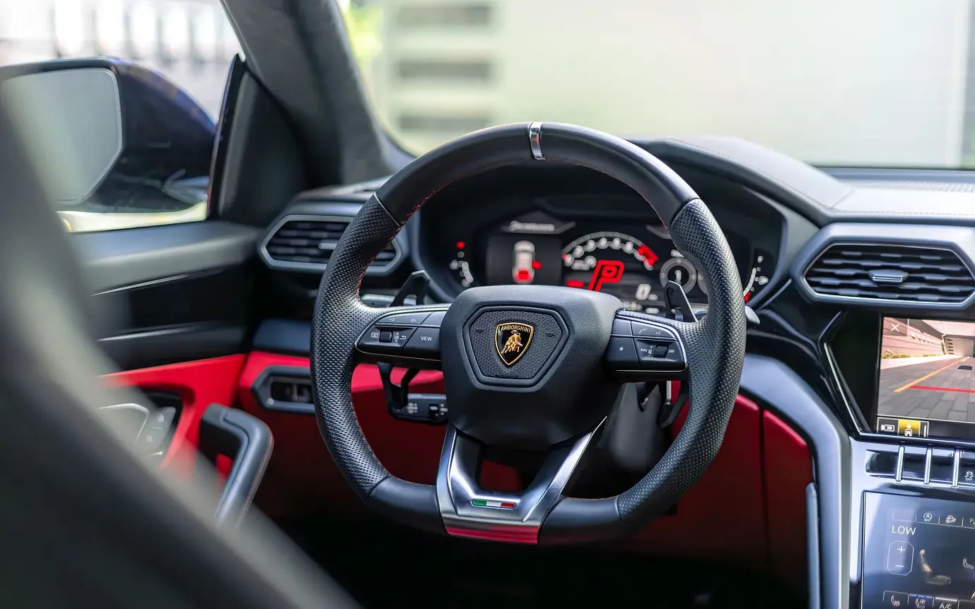 Interior of a luxury car, focusing on the steering wheel with Lamborghini logo, digital dashboard with red warning lights, and a screen displaying a rearview camera.
