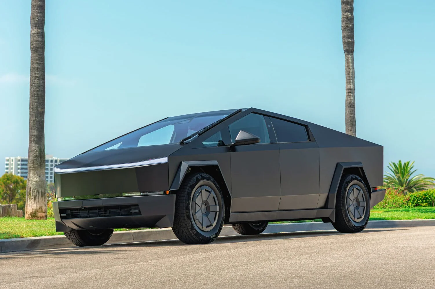 A sleek, futuristic black electric truck stands on a sunny street with palm trees in the background, showcasing its angular design.