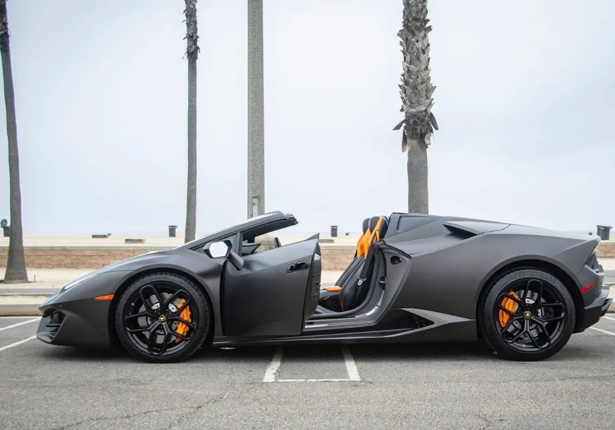 A matte gray Lamborghini Huracan Spyder with black wheels and orange brake calipers parked in an empty outdoor lot, with palm trees in the background and the convertible top open.