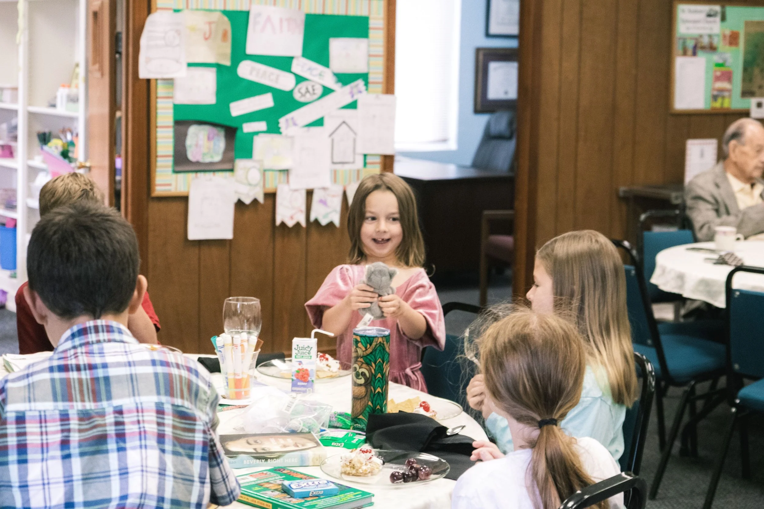 A young girl smiling and holding a stuffed animal at a birthday party with a table of snacks and drinks, surrounded by other children and adults in a decorated room.