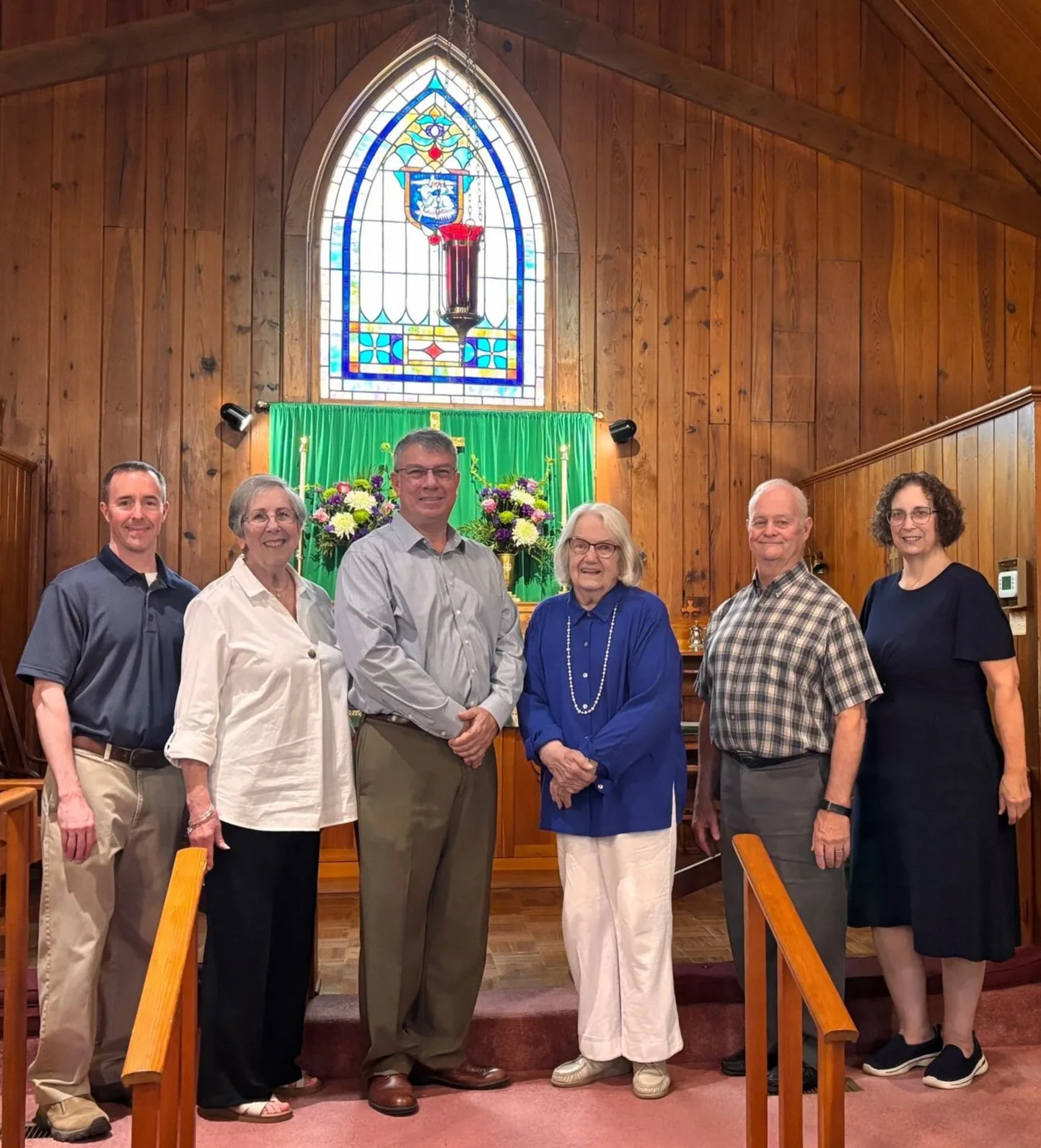 A group of seven people standing inside a church in front of a green altar with flowers and a large stained glass window.