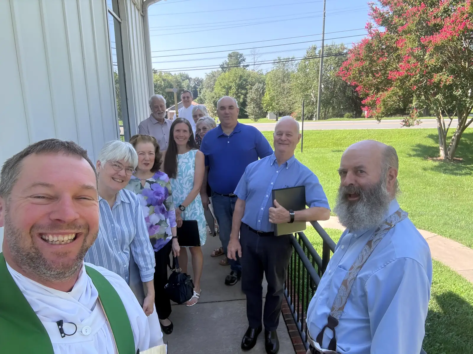 A group of smiling people taking a selfie outdoors on a sunny day near a building, with a grassy area and trees in the background.