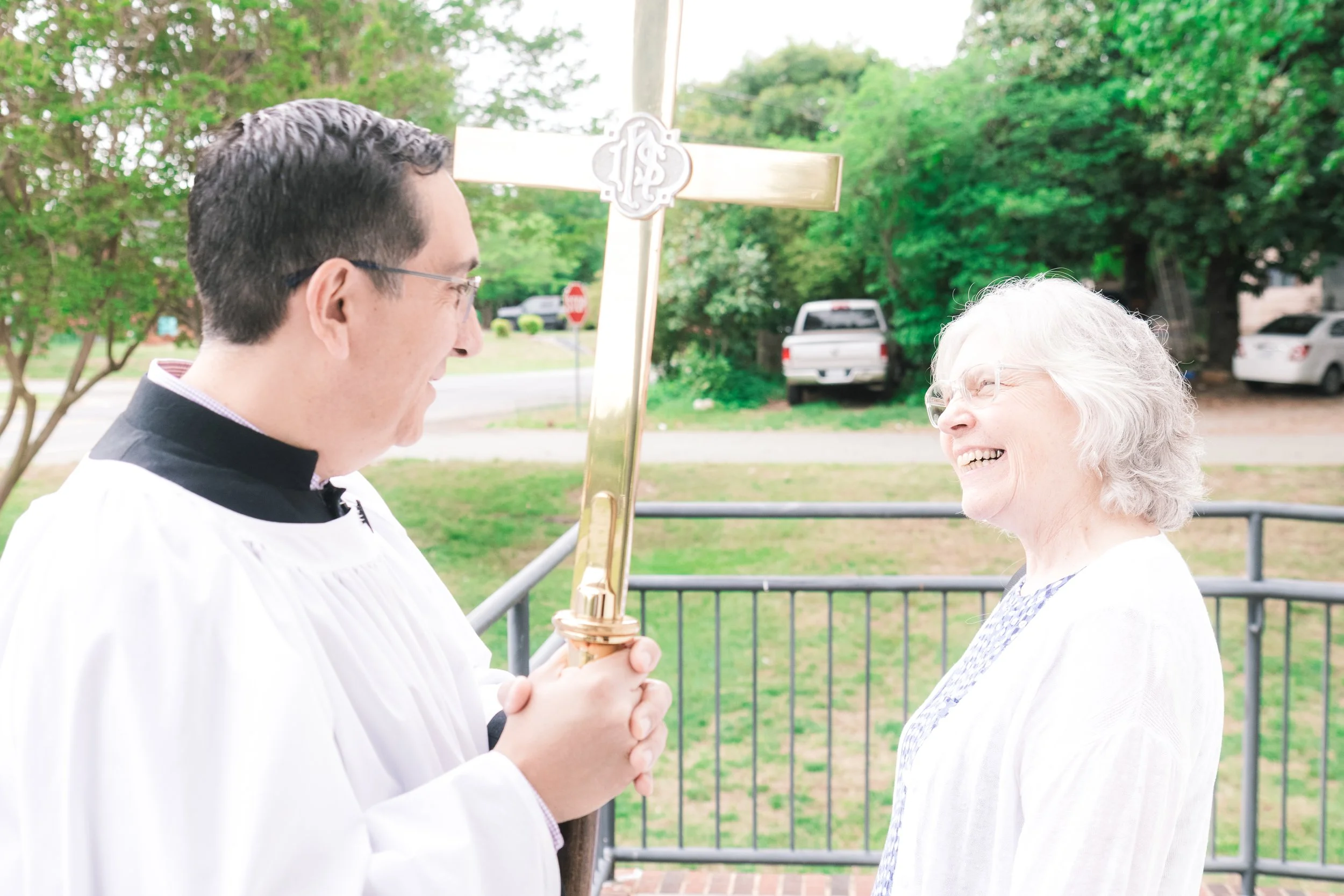 A priest holding a gold cross and clasping hands with an elderly woman, both smiling and making eye contact outdoors with trees in the background.