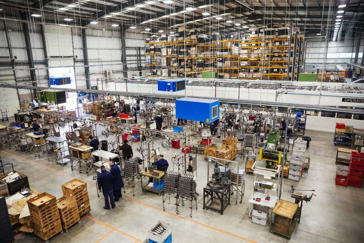 Inside a large industrial warehouse with workers at assembly stations, shelves filled with parts and equipment in the background.